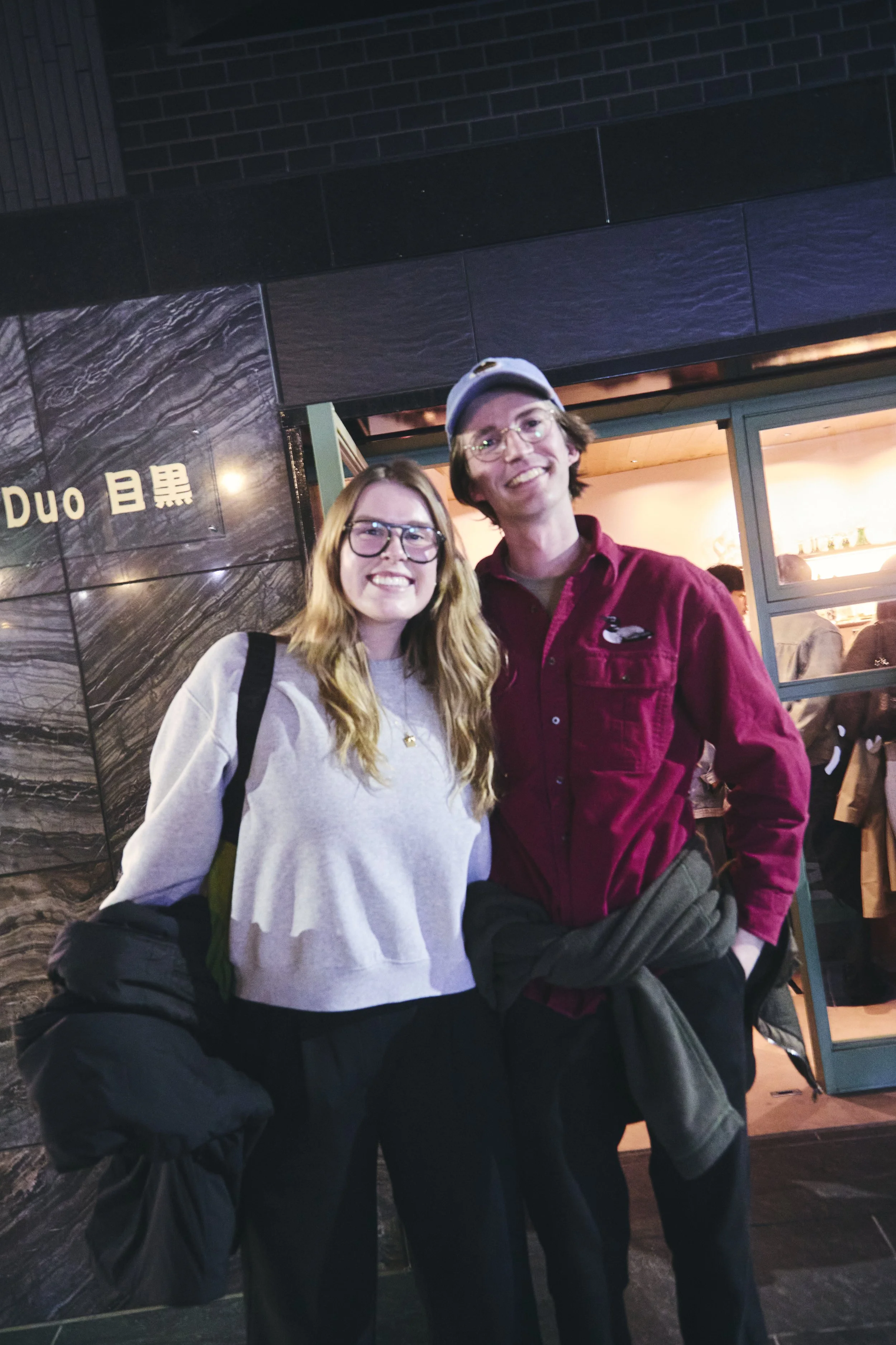 Two young adults, a woman with long red hair and glasses wearing a grey sweatshirt and a man with dark hair, glasses, and a backwards cap wearing a maroon shirt, pose together smiling in front of a black and grey wall with gold lettering and a window
