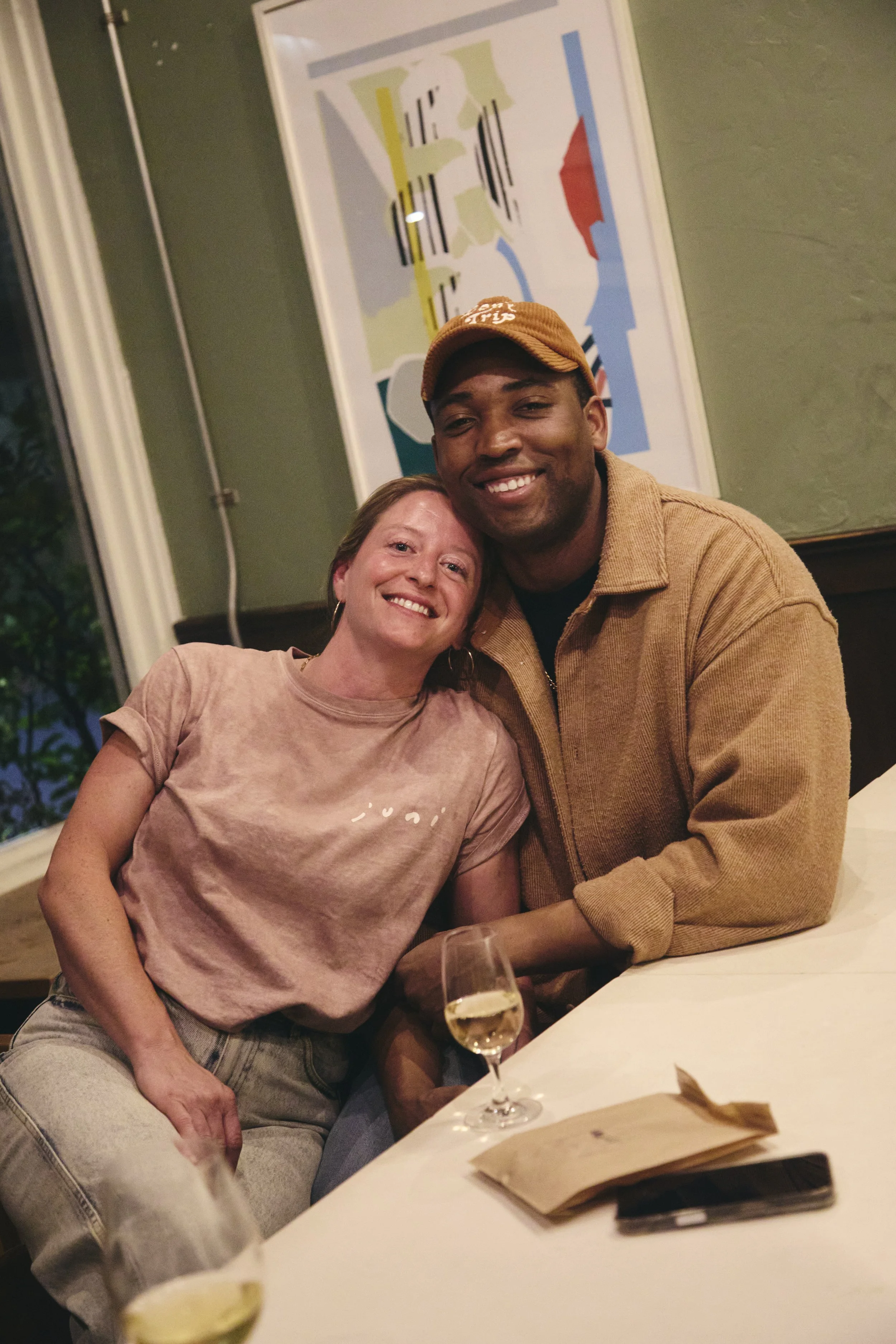 Two friends, a woman and a man, smiling and posing together at a restaurant, with a glass of white wine on the table and a colorful abstract painting on the wall behind them.