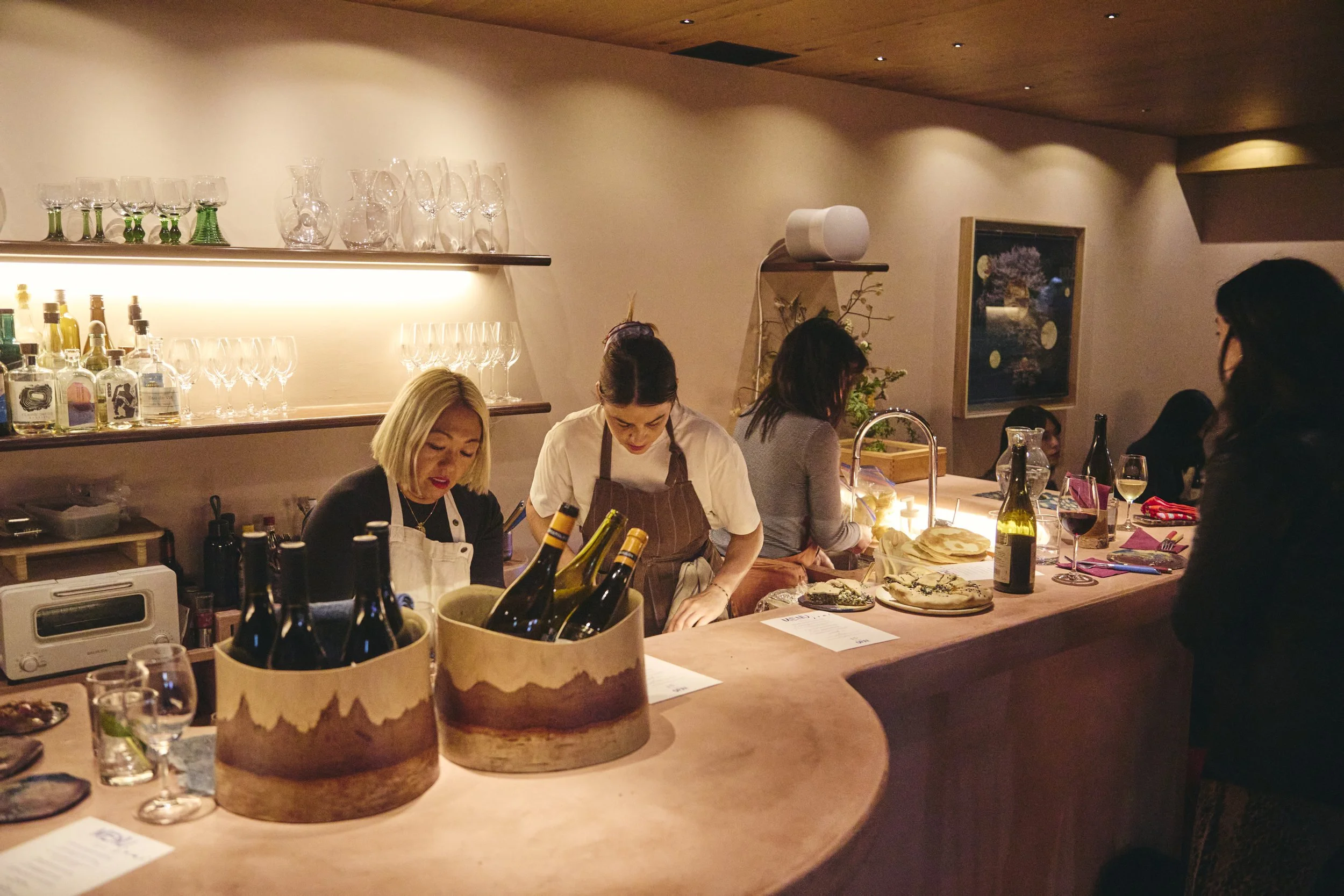 People working and preparing food behind a restaurant counter, with glasses, bottles, and food items visible.