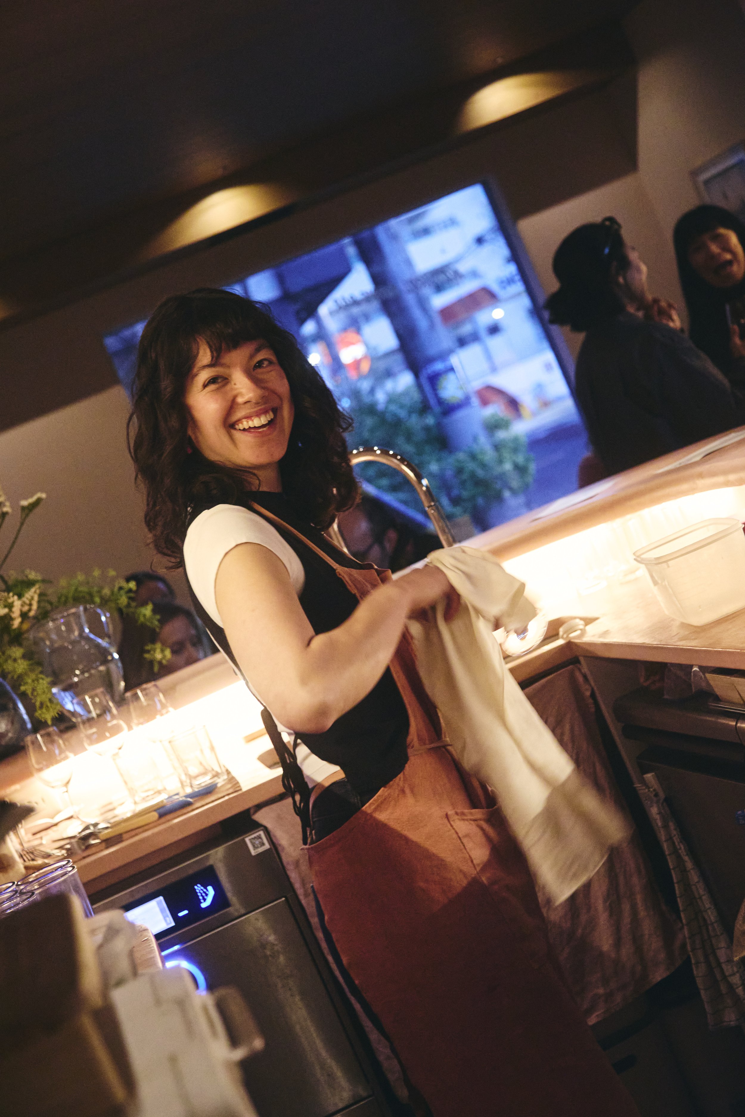 A woman smiling at a bar or restaurant, wearing a white t-shirt and a black apron, with several people in the background. The scene is dimly lit with a window showing an urban street outside.