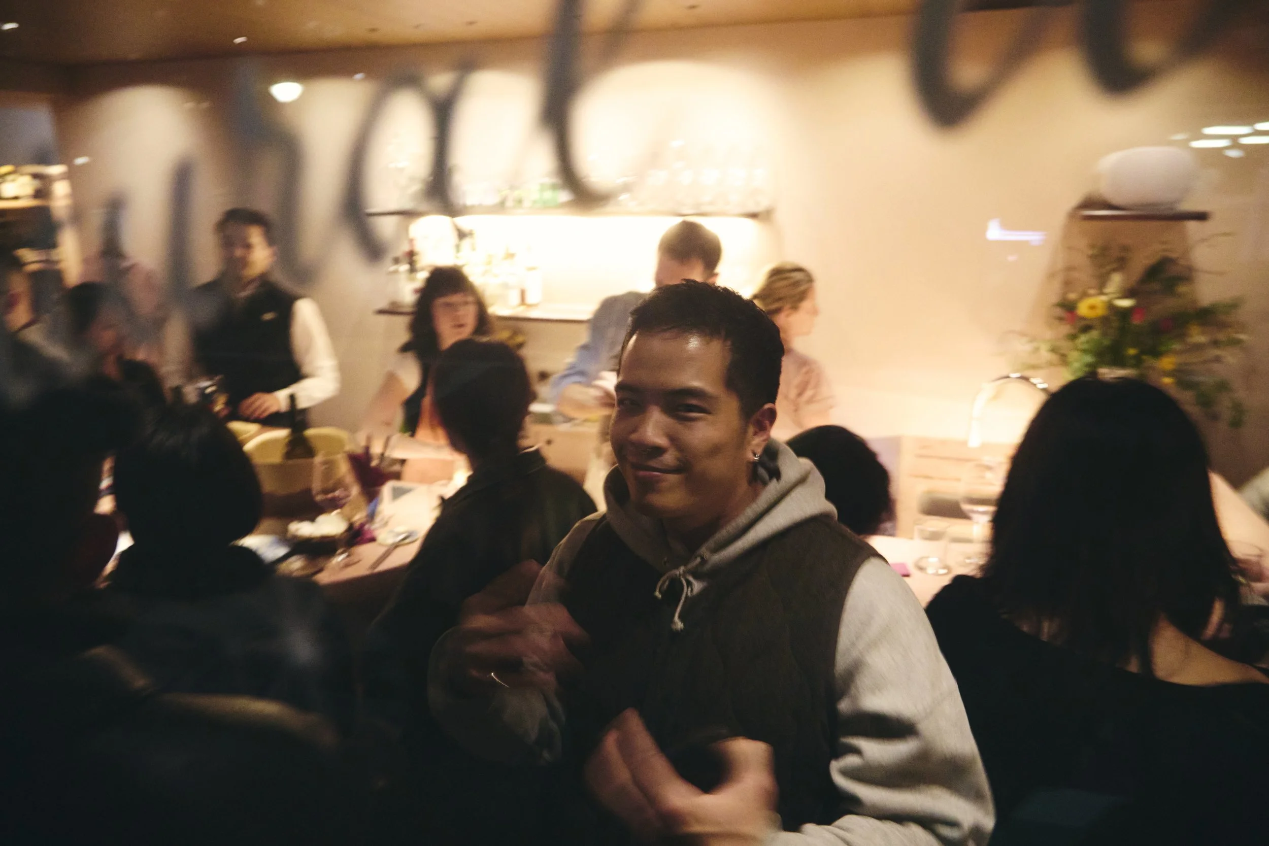 A group of people gathered in a cozy, warmly lit restaurant or bar, socializing and enjoying drinks. Three women and three men are visible, with one man smiling at the camera. The background features shelves with bottles and glasses, and a decorative