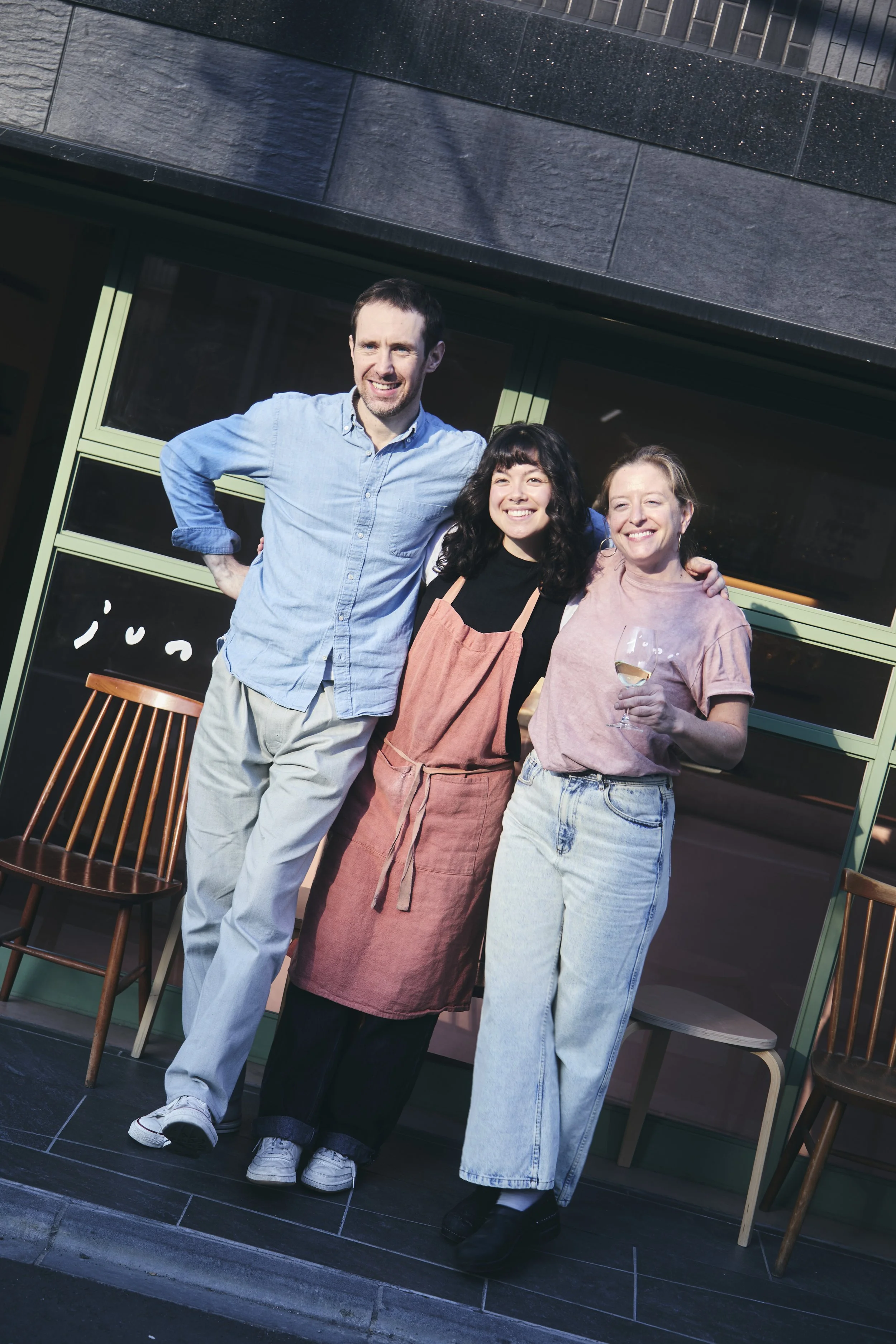 Three people posing outside a building, smiling at the camera. The man on the left wears a light blue shirt and light-colored pants, the woman in the middle wears a black top and coral dress, and the woman on the right wears a pink t-shirt and light 
