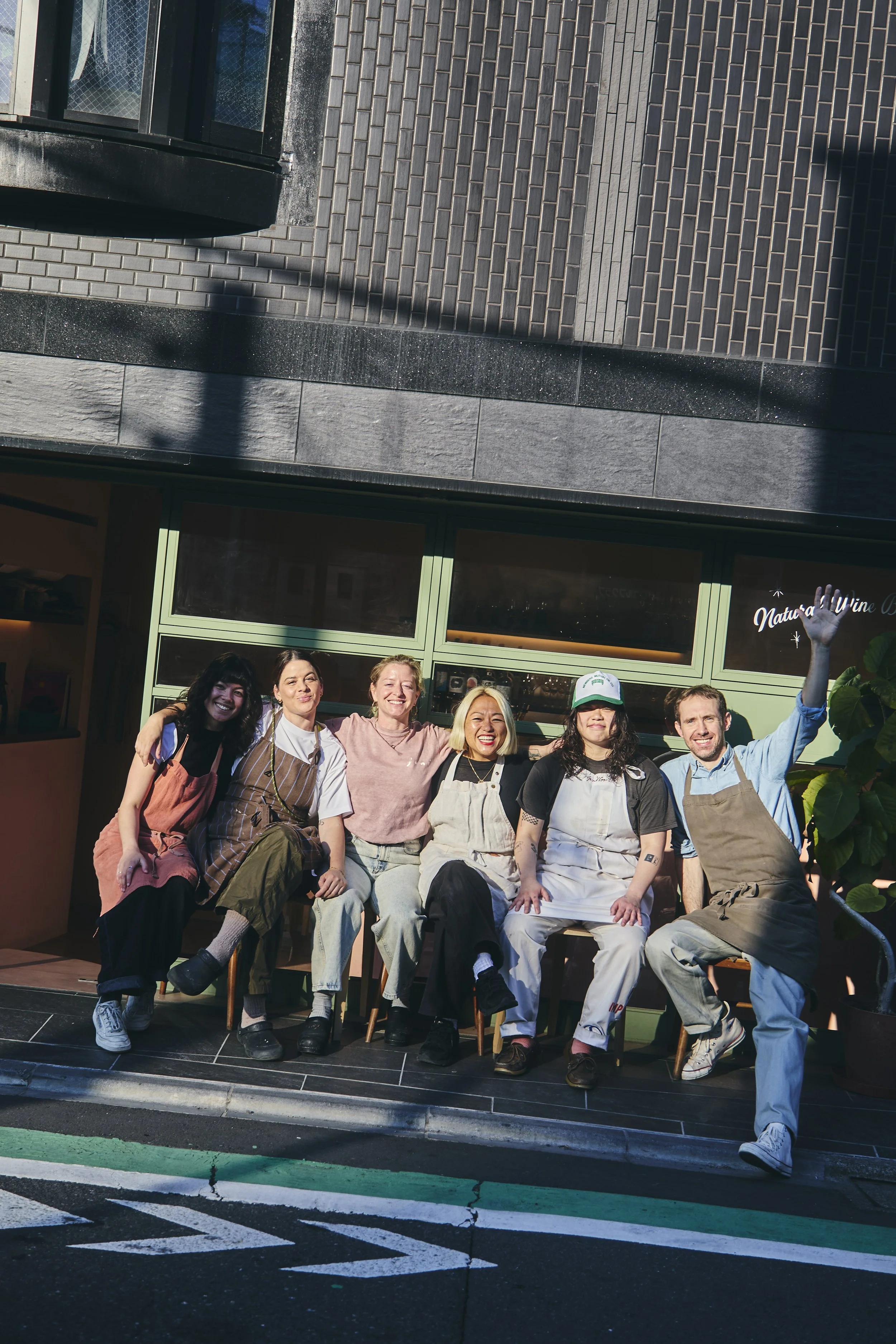 Group of seven diverse people sitting and standing outside a building, smiling and posing for a photo. Some are wearing aprons and casual clothing, indicating they may be involved in cooking or a similar activity. The building behind has large glass 