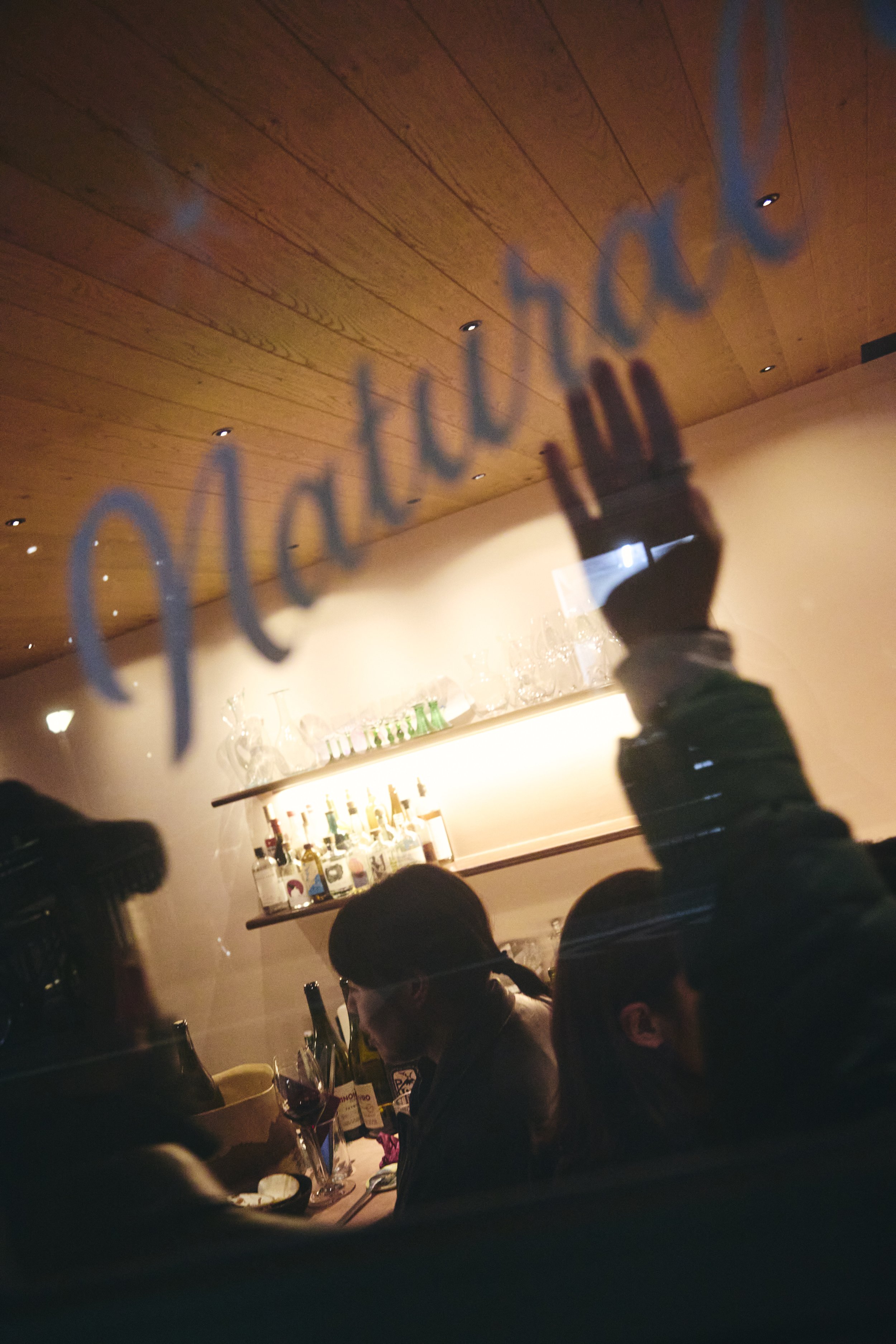 A reflection of a handwritten word 'welcome' on a glass surface in a dimly lit restaurant or bar, with several people sitting at tables, some drinking wine, and bottles of alcohol on a shelf in the background.