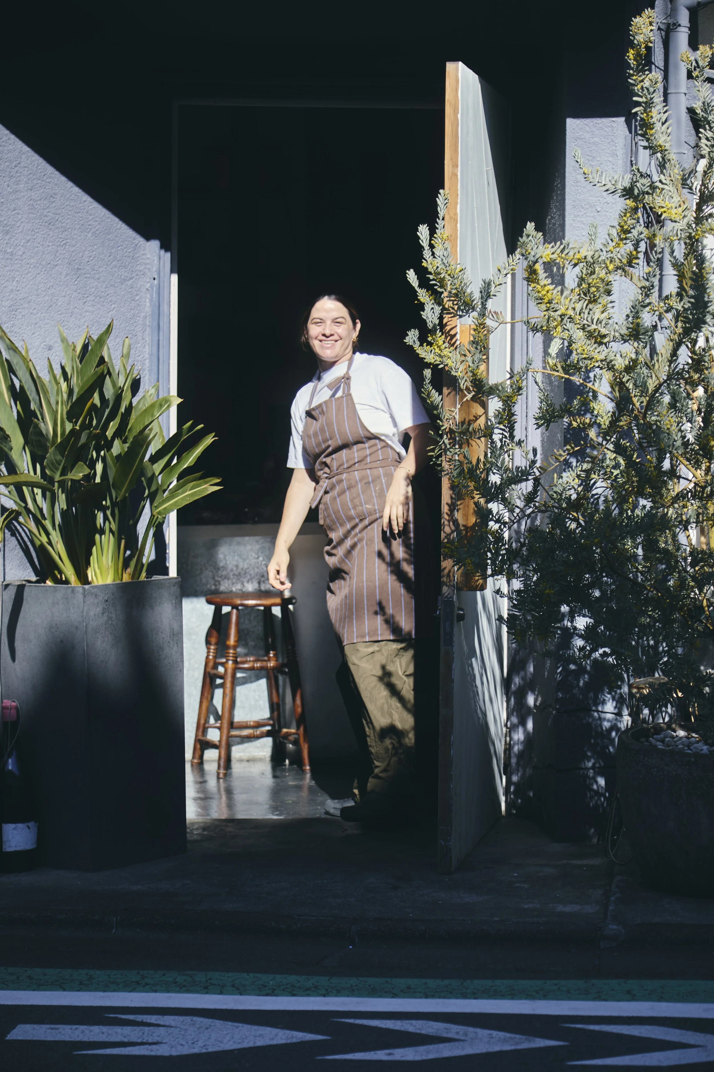 A smiling woman wearing a white shirt and a striped apron standing at the entrance of a dark room, partially outside on a sunny day, with large potted plants nearby.