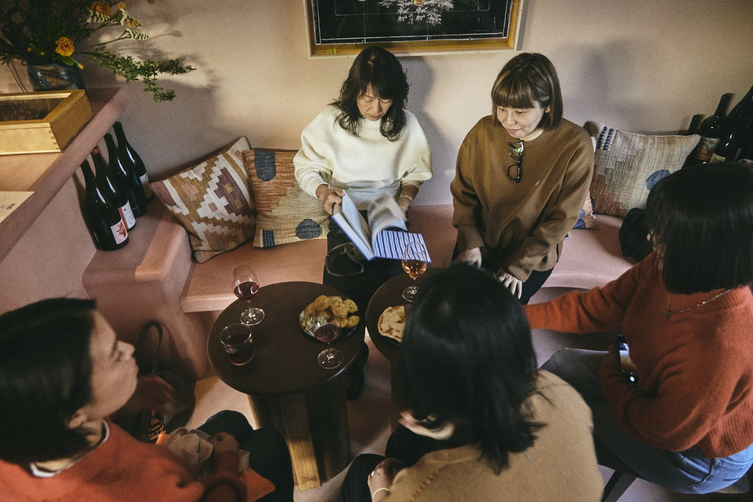 Group of people gathered around a table in a cozy room, enjoying food and drinks, with some women looking at a book or menu.