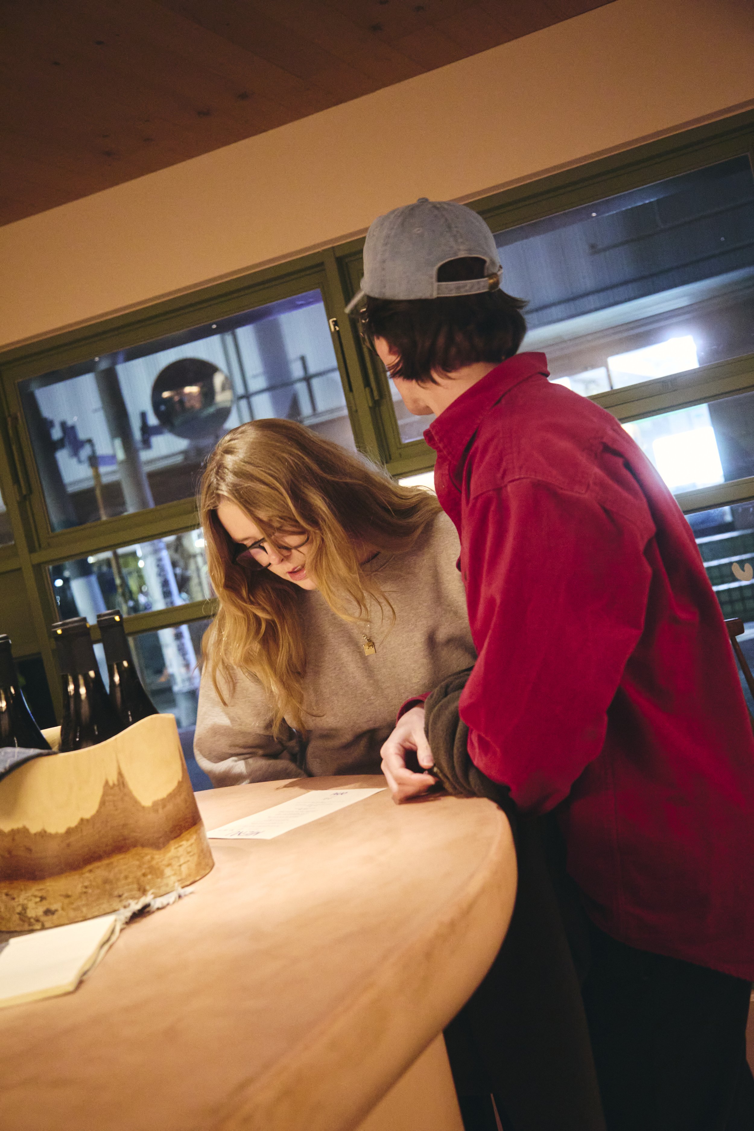 A woman with long blonde hair, glasses, and a gray sweater is sitting at a wooden table, looking down at a paper. A man with dark hair, wearing a gray cap and a red jacket, is standing next to her, also looking at the paper. The scene appears to be i