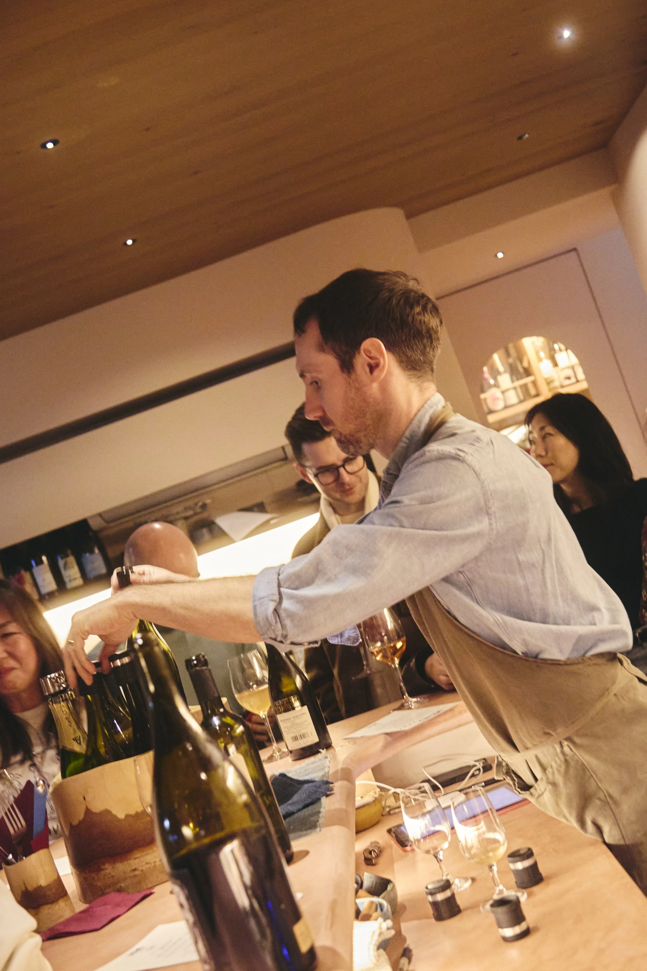 A man serving wine at a restaurant table with other people and wine bottles in the background.