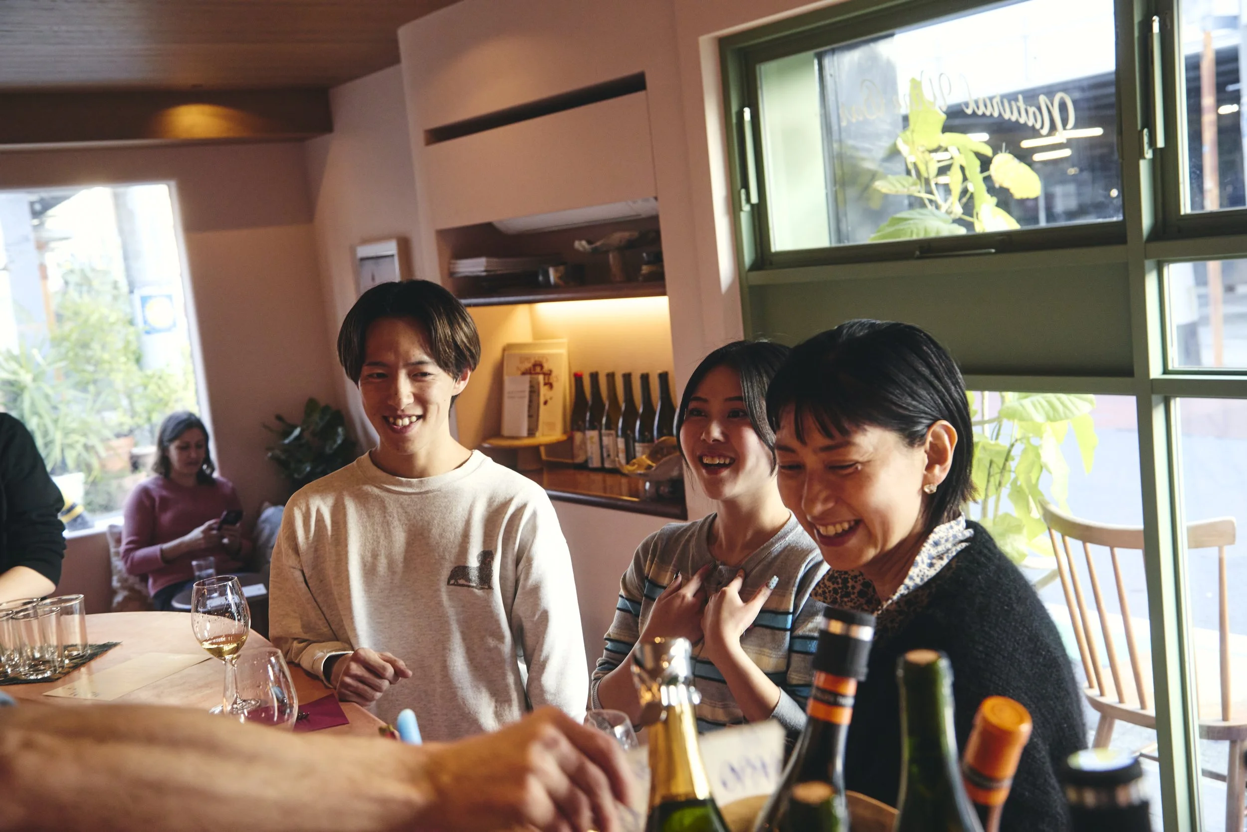A group of people smiling and enjoying drinks at a restaurant or cafe with sunlight coming through large windows.
