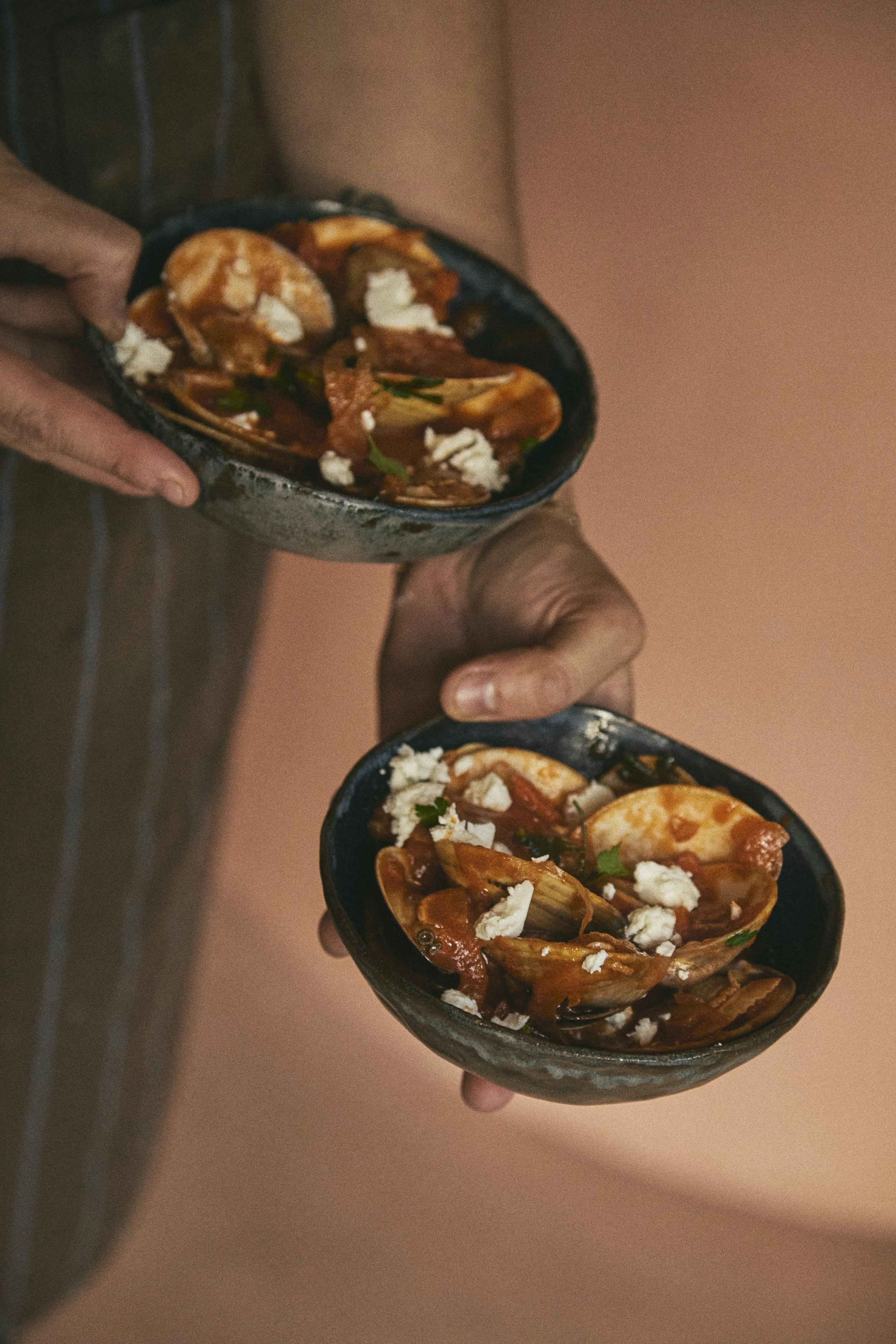 Two hands holding black bowls filled with chili topped with crumbled cheese and herbs.