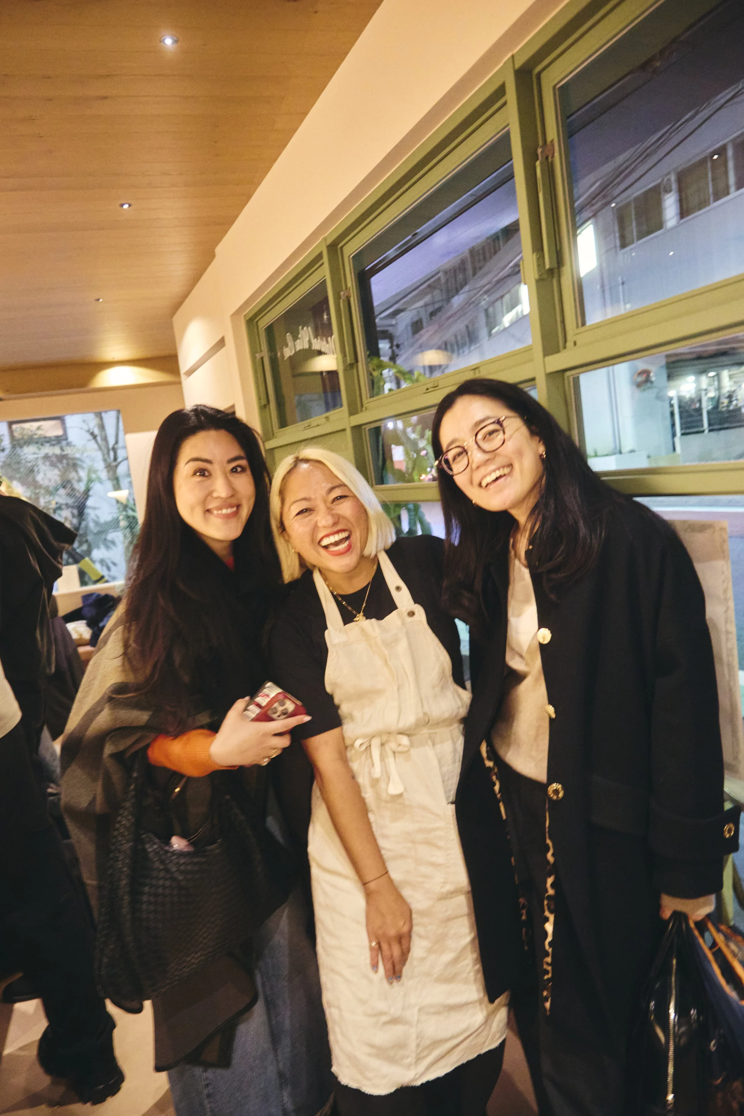 Three women smiling and posing together indoors near a window with green window frames. The woman in the middle is wearing a beige apron, and the other two women are wearing dark jackets. They all appear happy and cheerful.