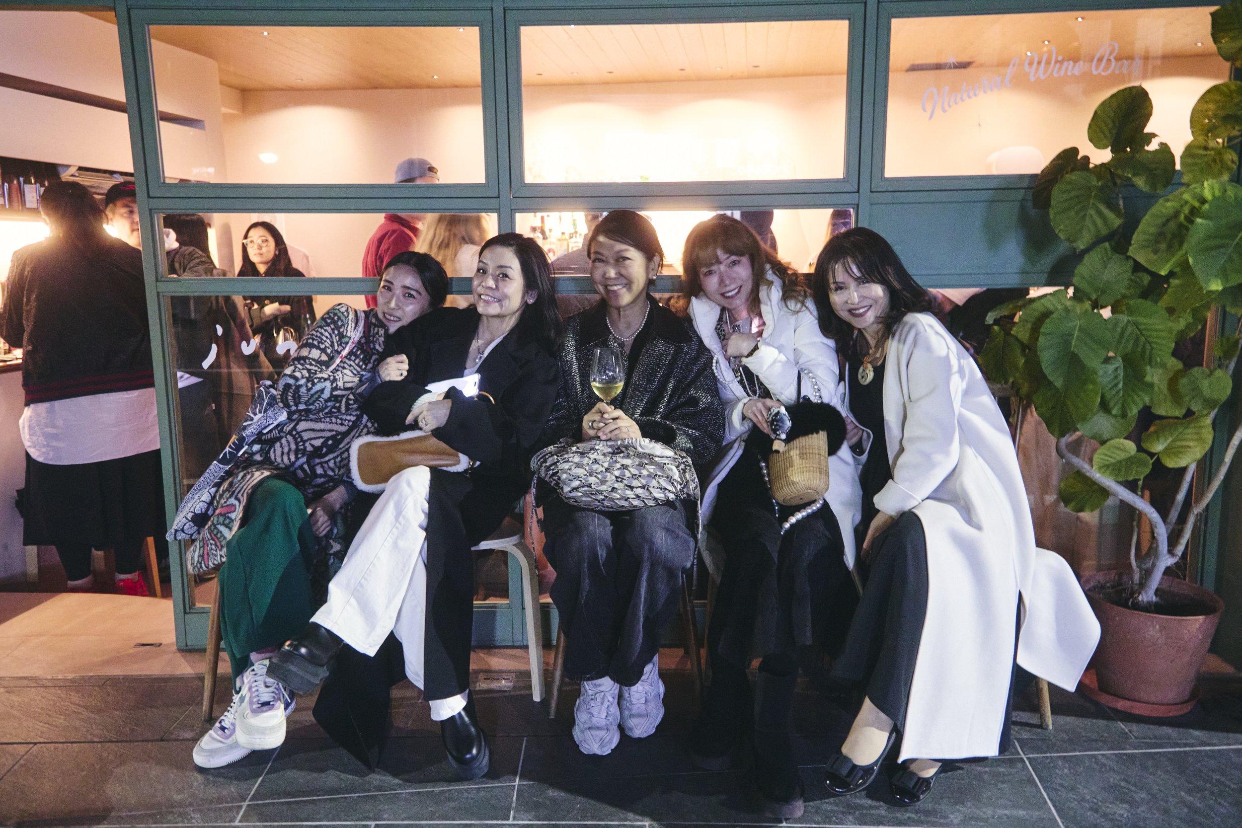 Group of five women sitting together at a restaurant table, smiling at the camera, with other guests and a bar in the background.