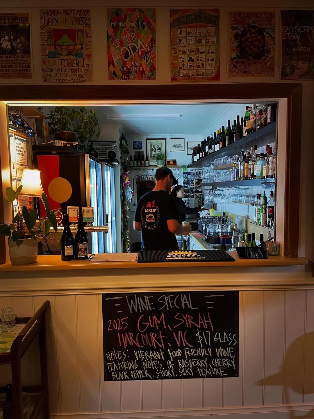 View of a cozy bar with two bartenders behind the counter, shelves stocked with wine bottles and glasses, and a blackboard sign advertising a wine special featuring GUM, SIRAH, HARCOURT, and VIC, priced at $17 per glass, with notes about the wine's v