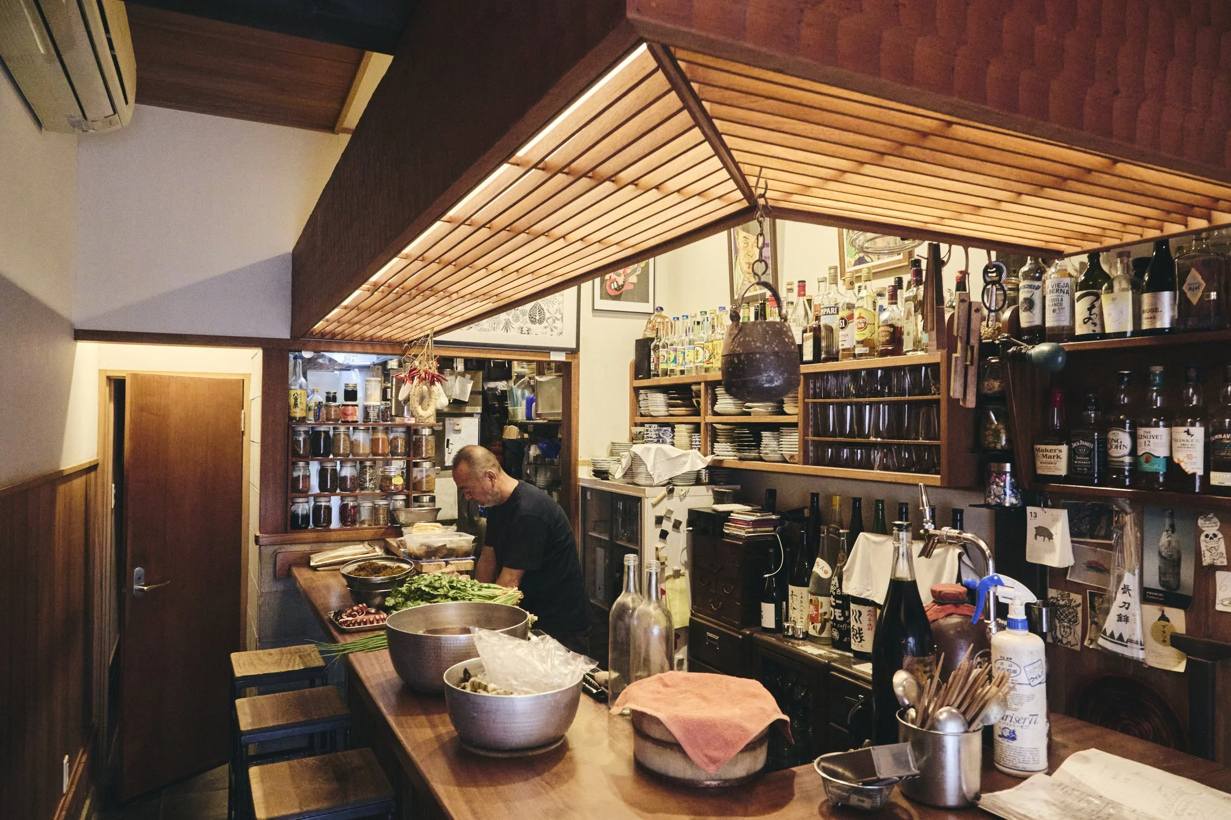 Interior of a cozy restaurant with a wood-paneled ceiling, shelves of bottles and plates, and a chef preparing food at a counter.
