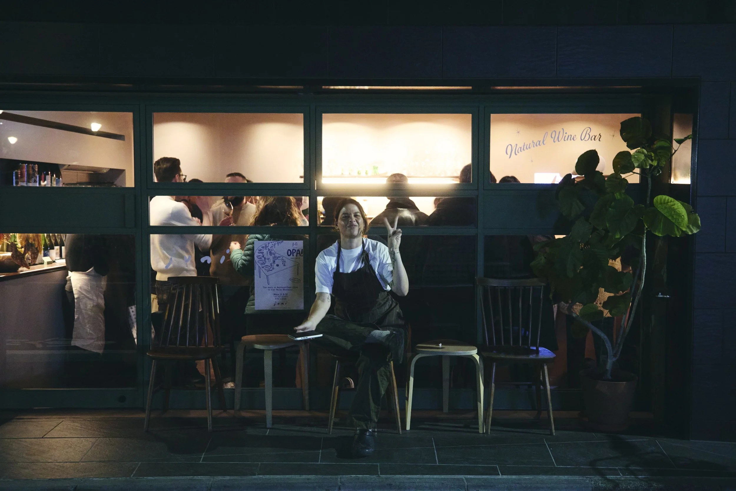 A woman sitting outside a wine bar making a peace sign with her fingers, with a window showing people inside a crowded wine bar.