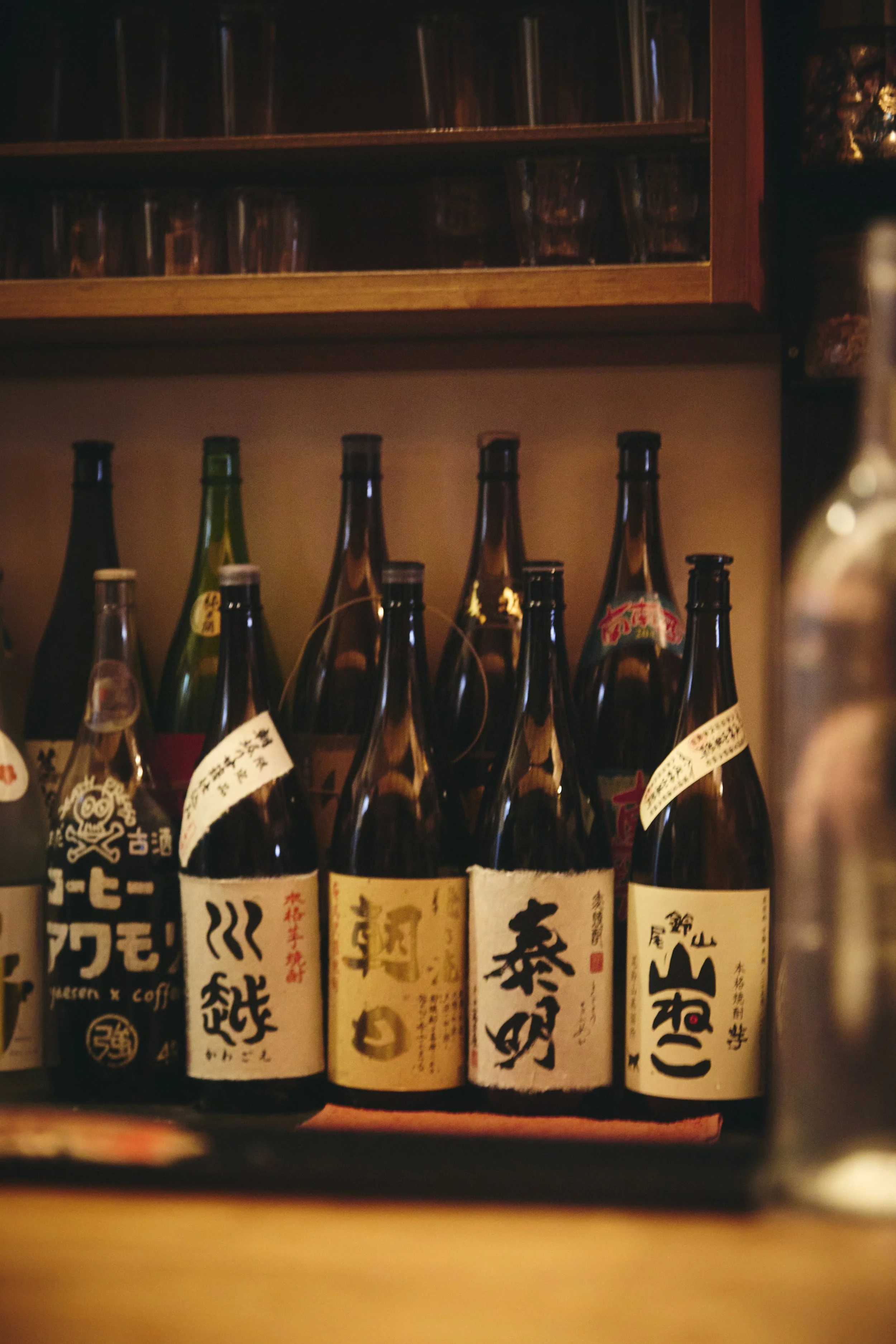 Shelf with several bottles of Japanese sake or similar alcoholic beverages with Japanese labels in a dimly lit setting.