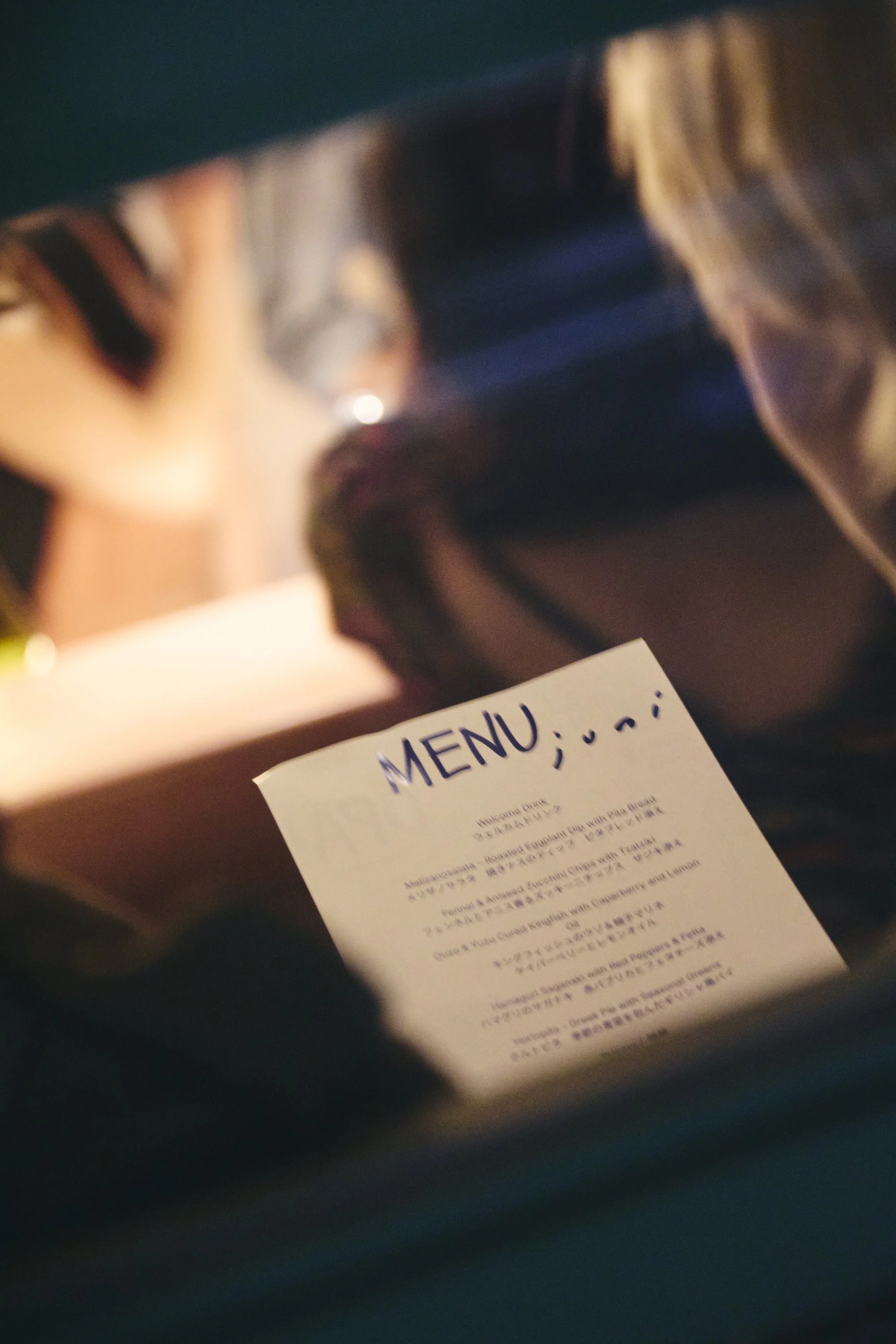 A blurry image of a menu and a person's hand holding a pen, with a dark background and some visible lighting reflections.