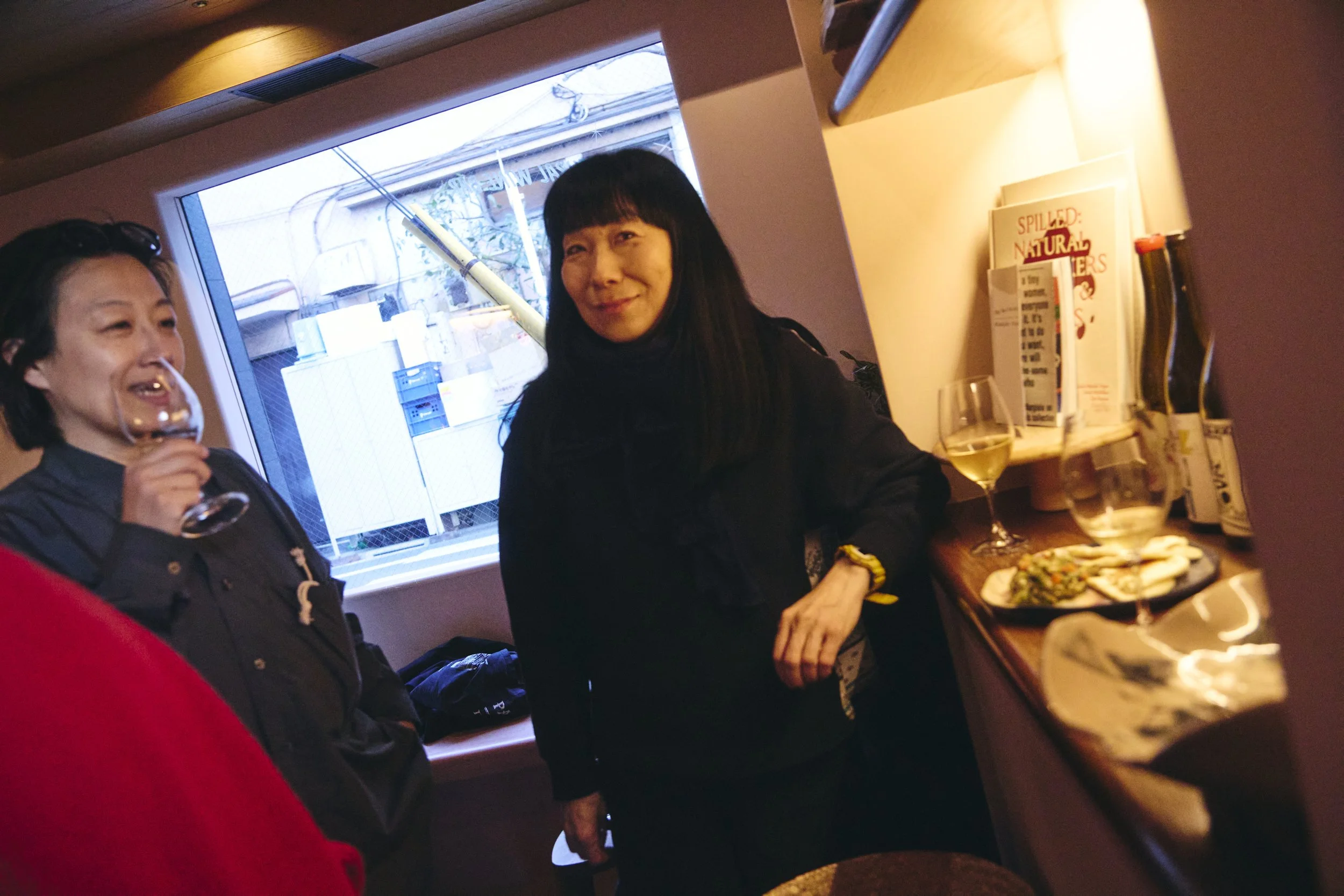 Two women standing near a window, one holding a wine glass, inside a room with a bookshelf and trays of food on a counter.
