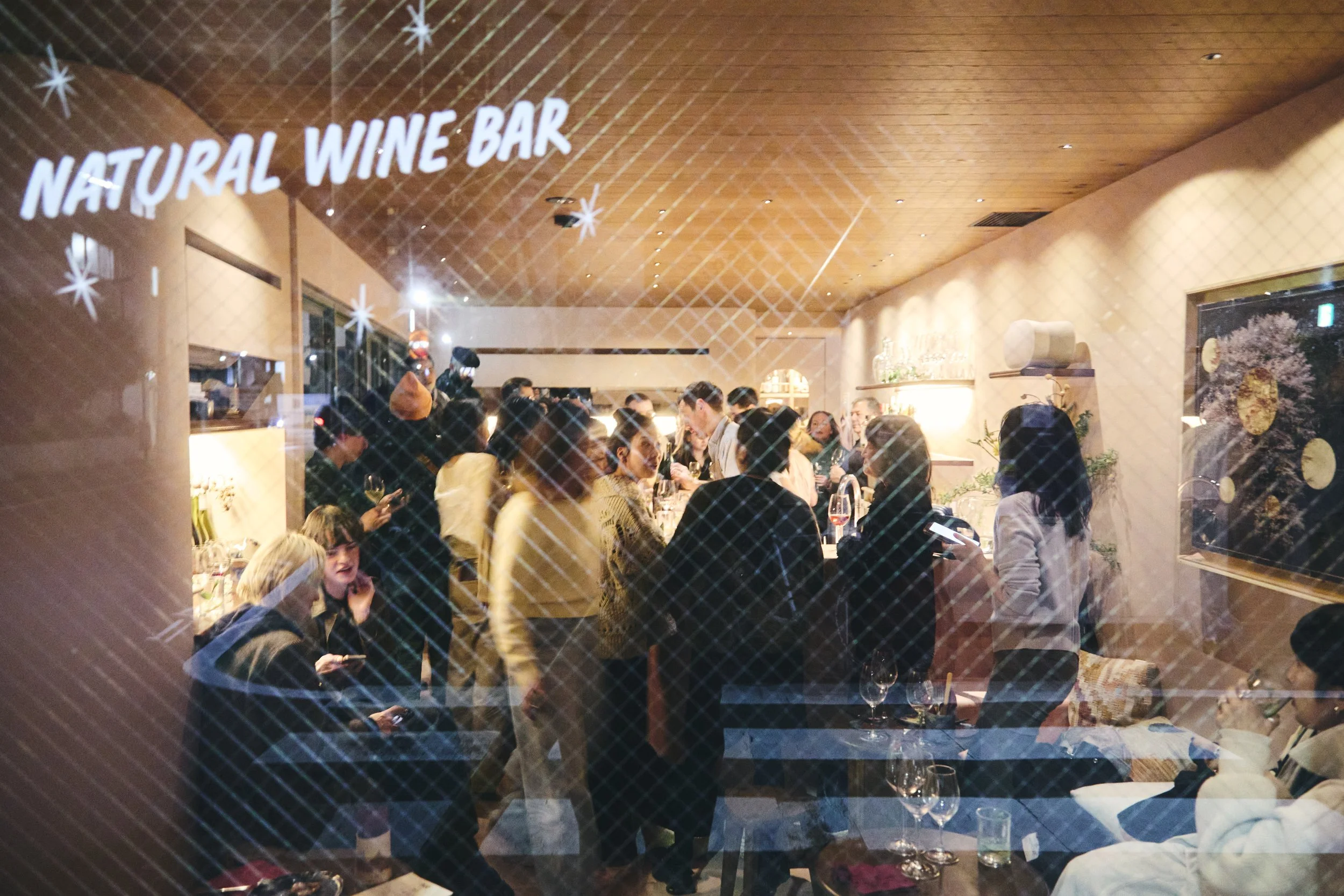 Inside a restaurant or wine bar with people dining and socializing; the view is through a textured or patterned glass window with the words "Natural Wine Bar" projected on it.