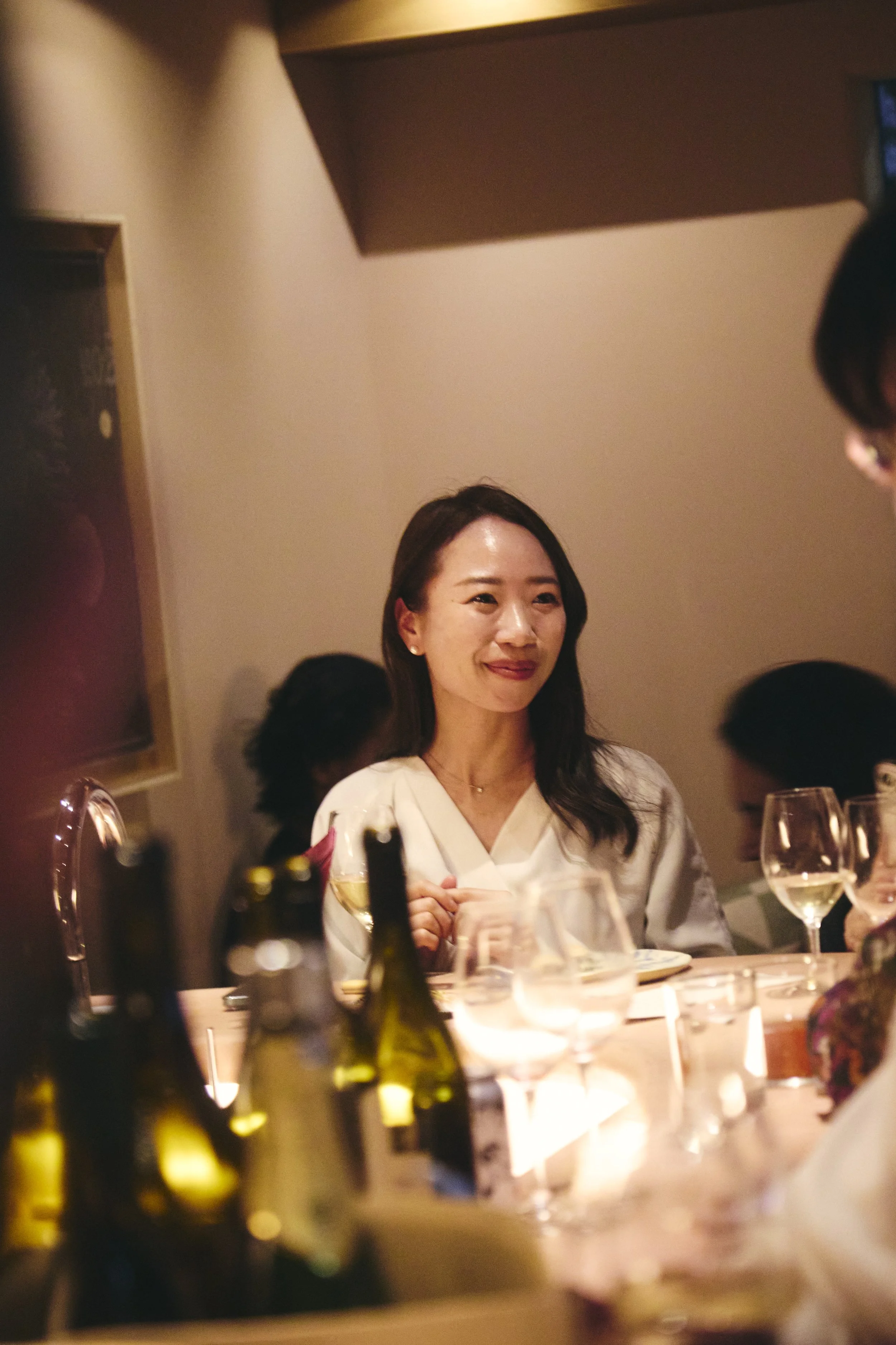 A woman with dark hair smiling during a dinner party, surrounded by glasses of wine and bottles on the table, with ambient lighting and blurry background.