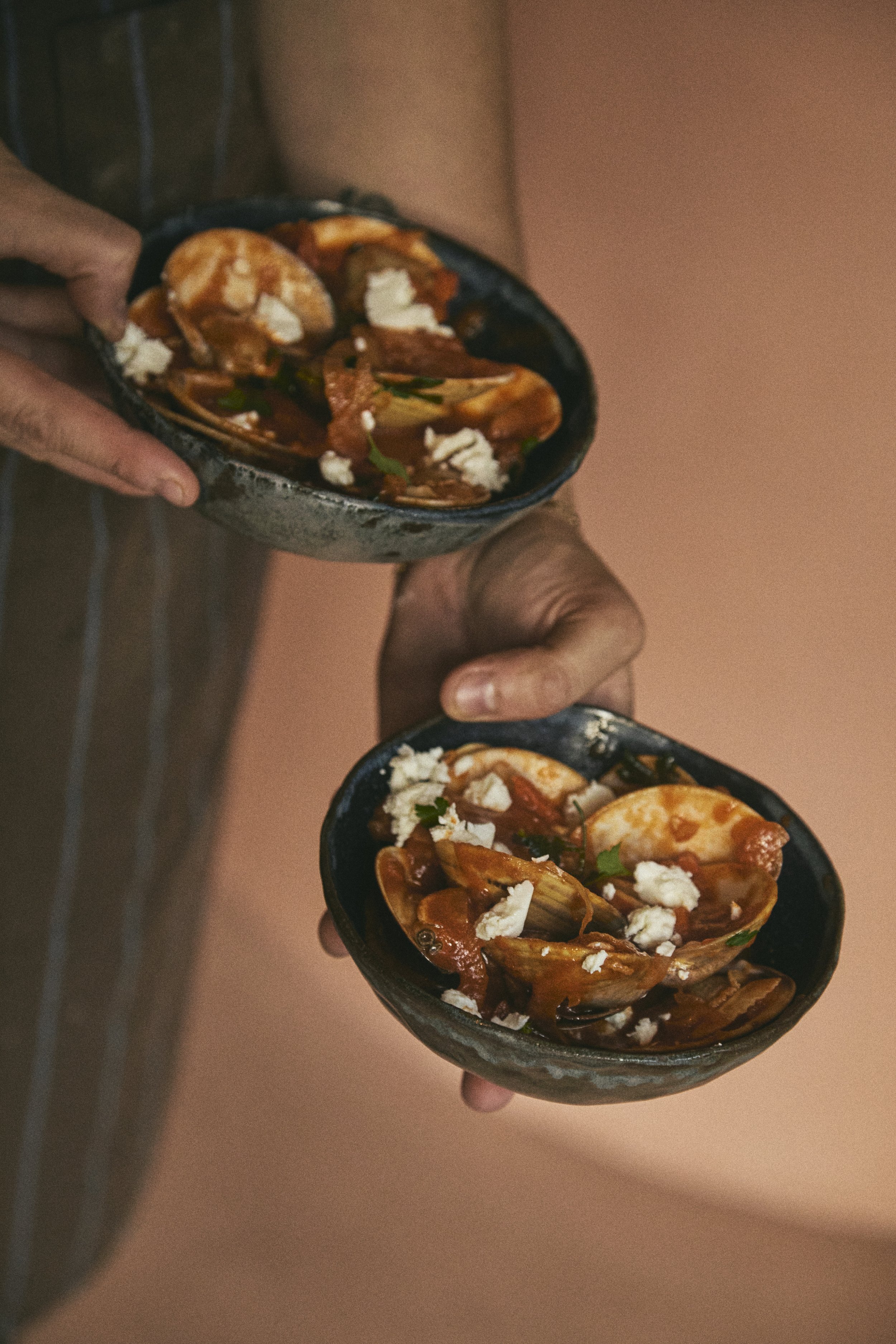 Close-up of two hands holding small black bowls filled with Mexican-style shrimp cocktails garnished with feta cheese and chopped herbs.