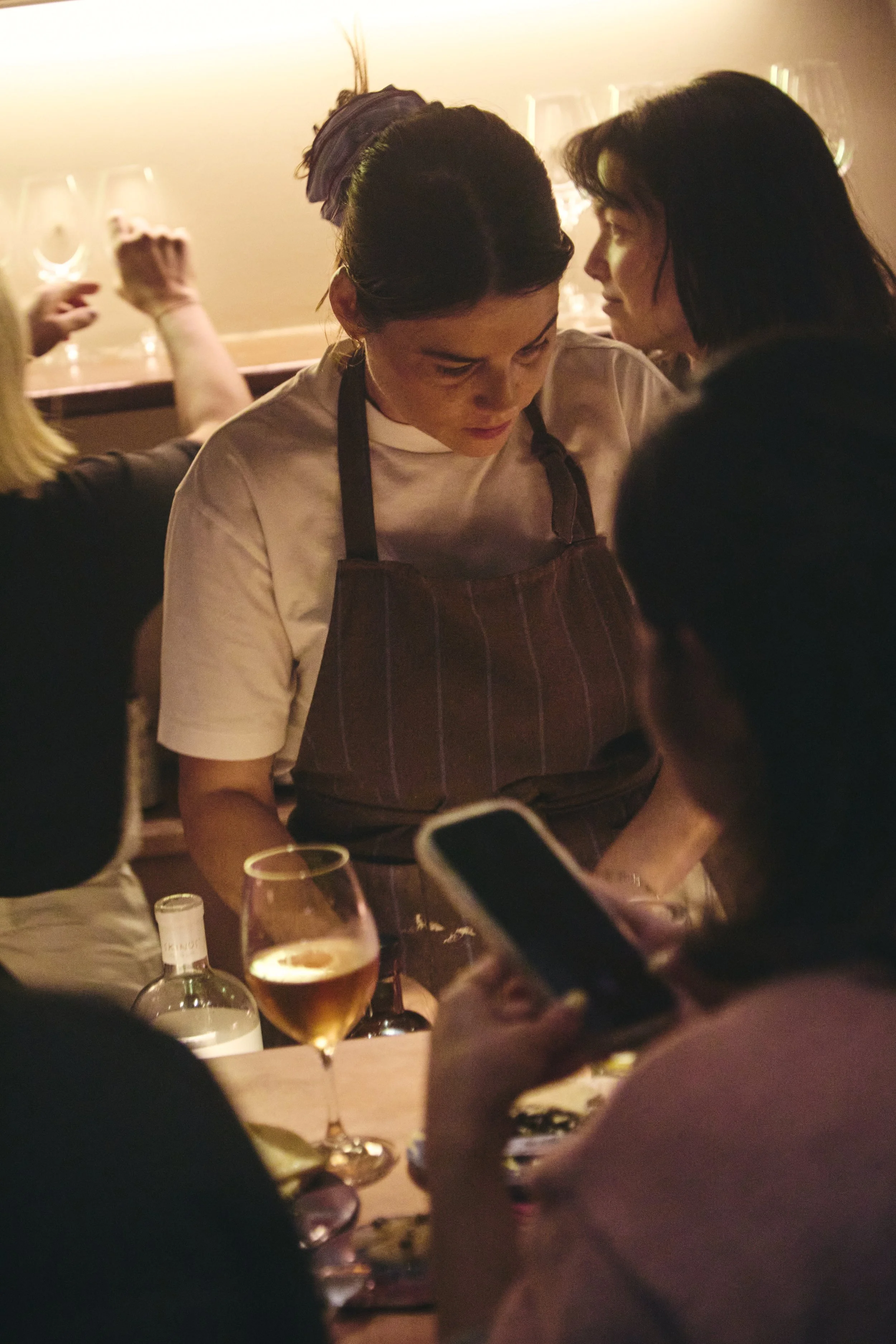 Two women in conversation at a dimly lit restaurant, with glasses of wine on the table and blurred figures in the background.