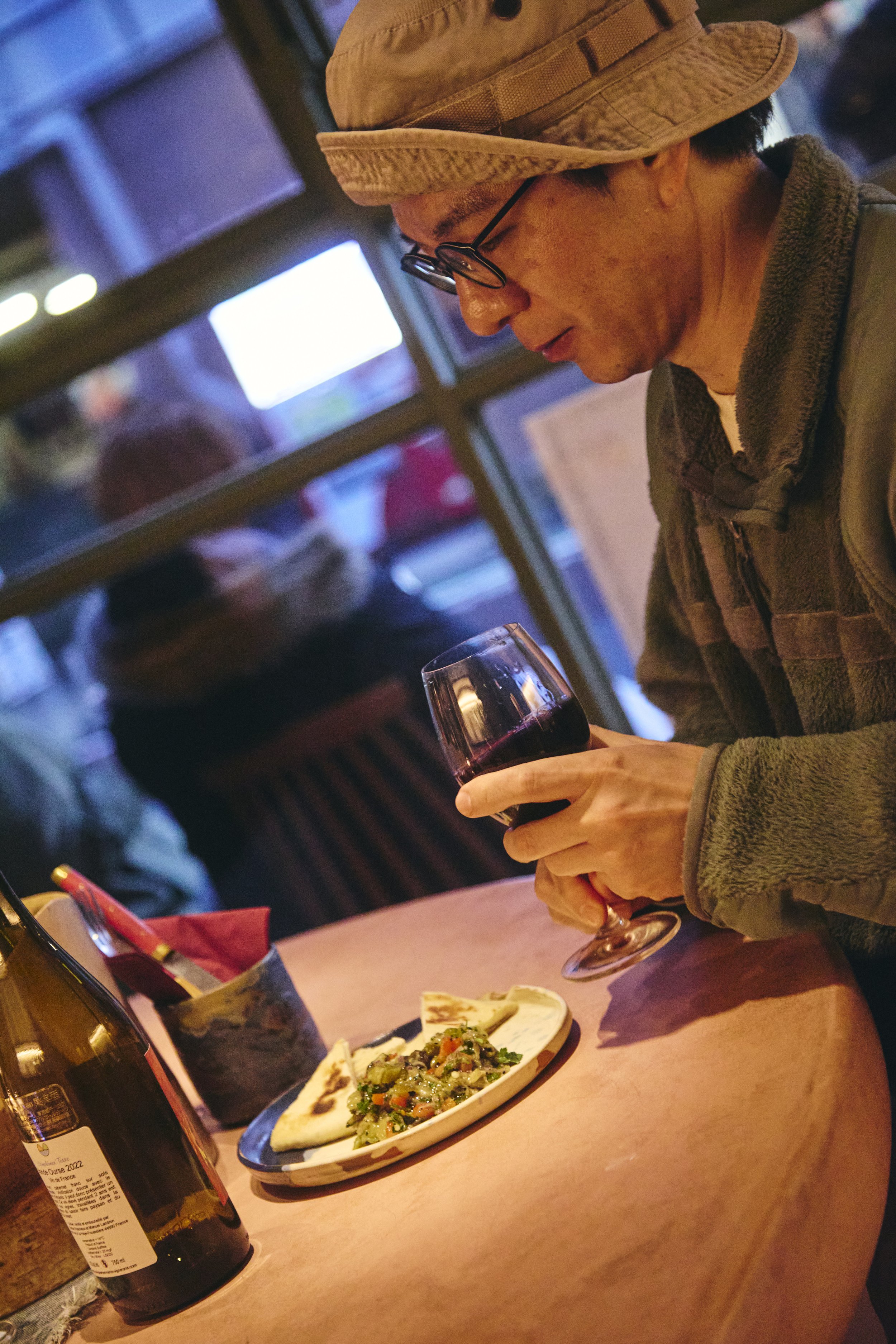 A man wearing glasses, a beige bucket hat, and a gray jacket is sitting at a restaurant table, holding a glass of red wine and looking down. On the table, there is a plate of food with vegetables and flatbread, a bottle of wine, and other dining item