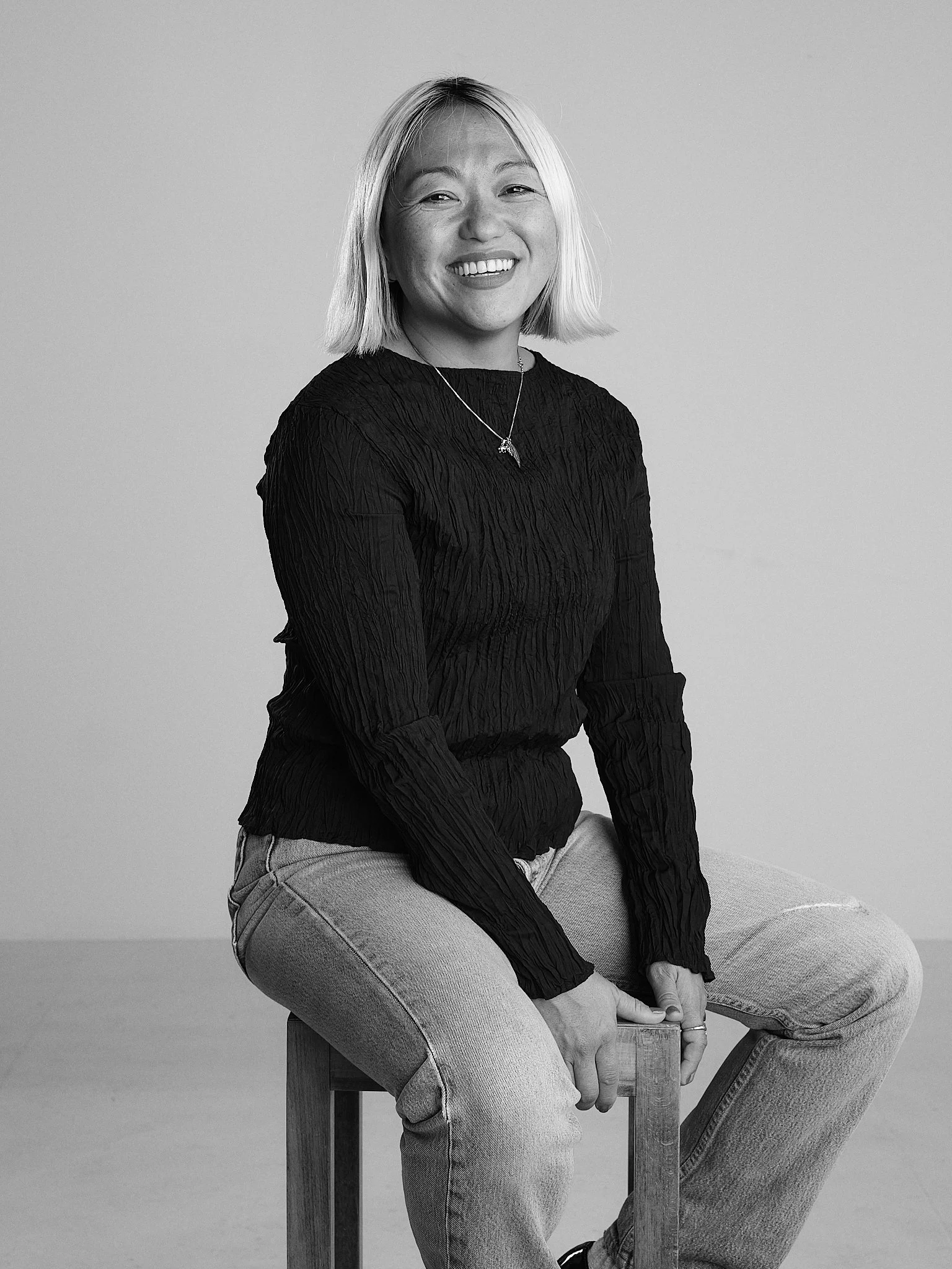 A woman sitting on a wooden stool, smiling, wearing a black textured long-sleeve top, light-colored jeans, and a necklace with a pendant. The photo is in black and white.