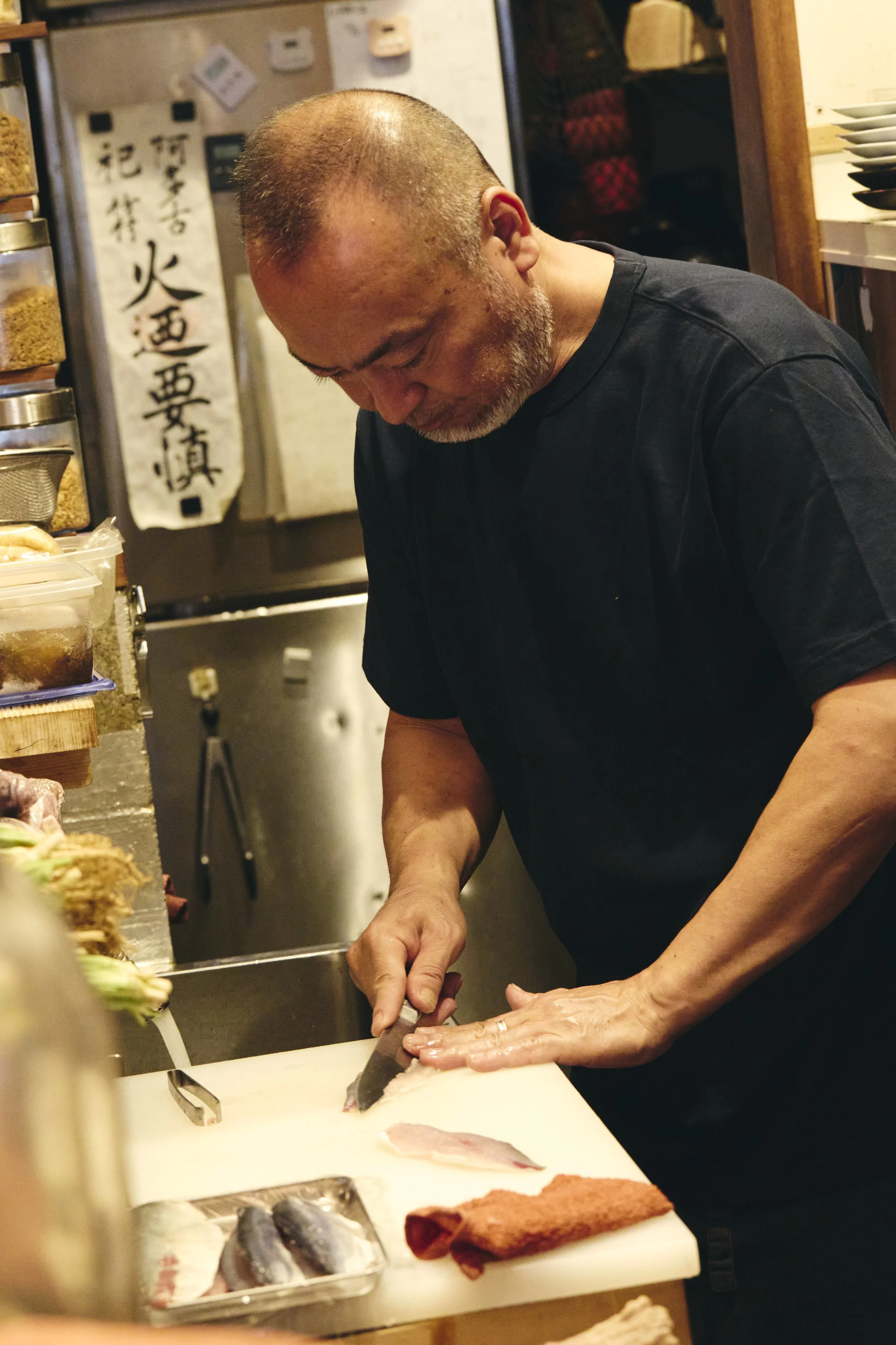 A man with a shaved head and gray beard is slicing fish on a cutting board in a kitchen. Various jars and plates are visible behind him, along with kitchen utensils and ingredients.