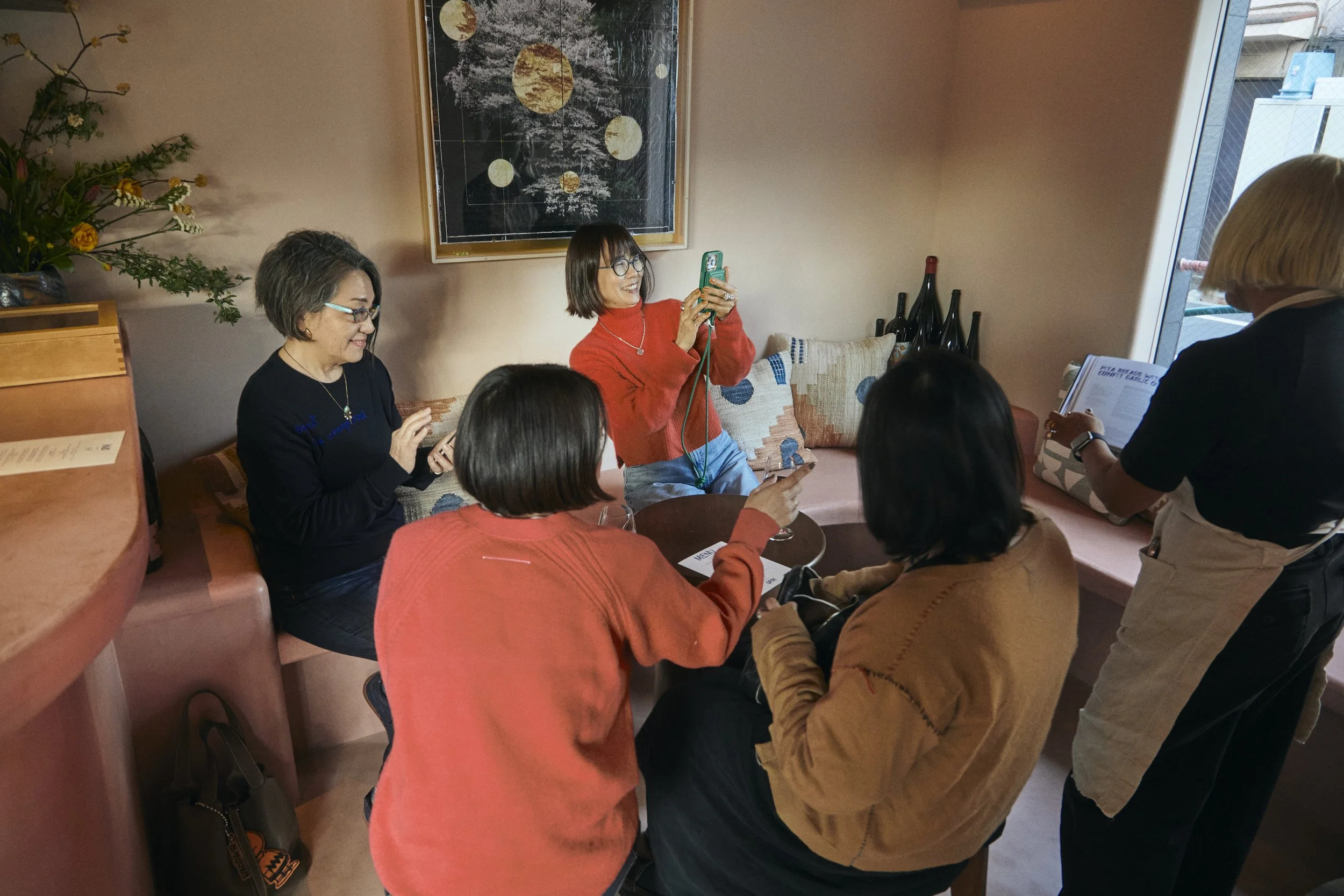 A group of six women sitting and standing around a pink cushioned booth in a restaurant or cafe, laughing and looking at their phones. One woman stands while holding her phone, taking a photo or video of the group. The background has a framed map or 