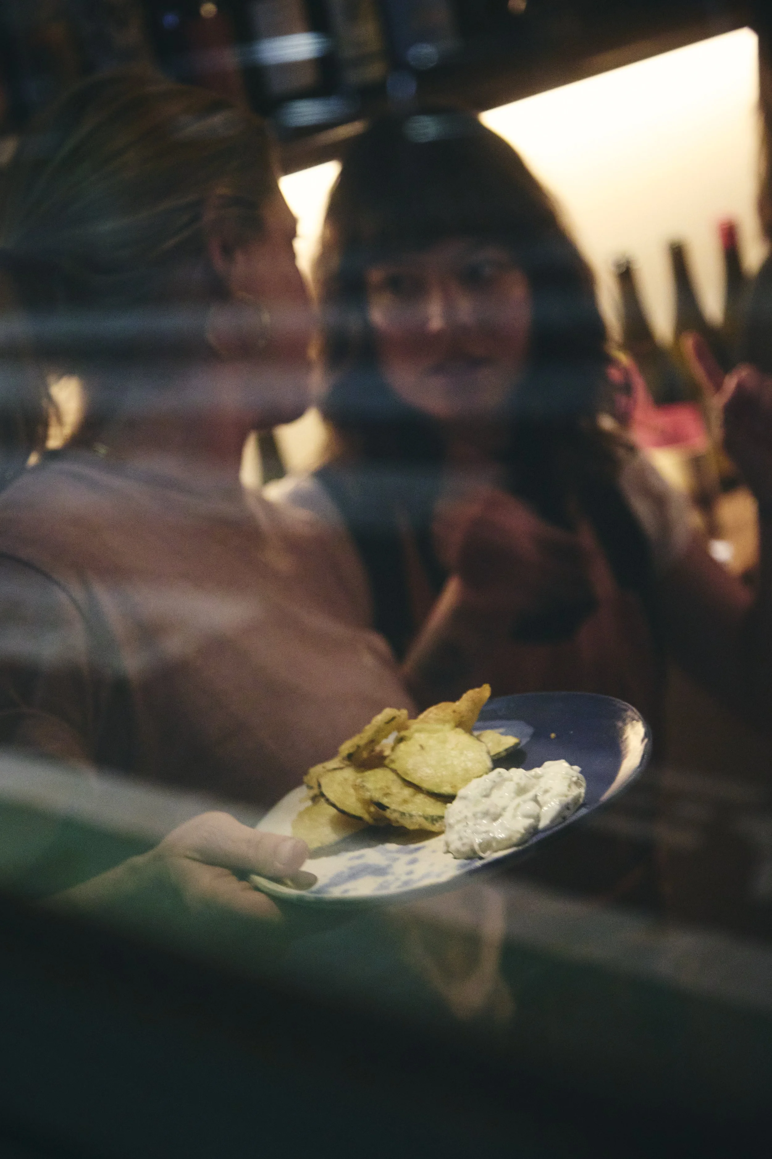A person holding a plate of food with potato chips and a creamy dip, viewed through a glass window with a blurred background of two women in conversation.