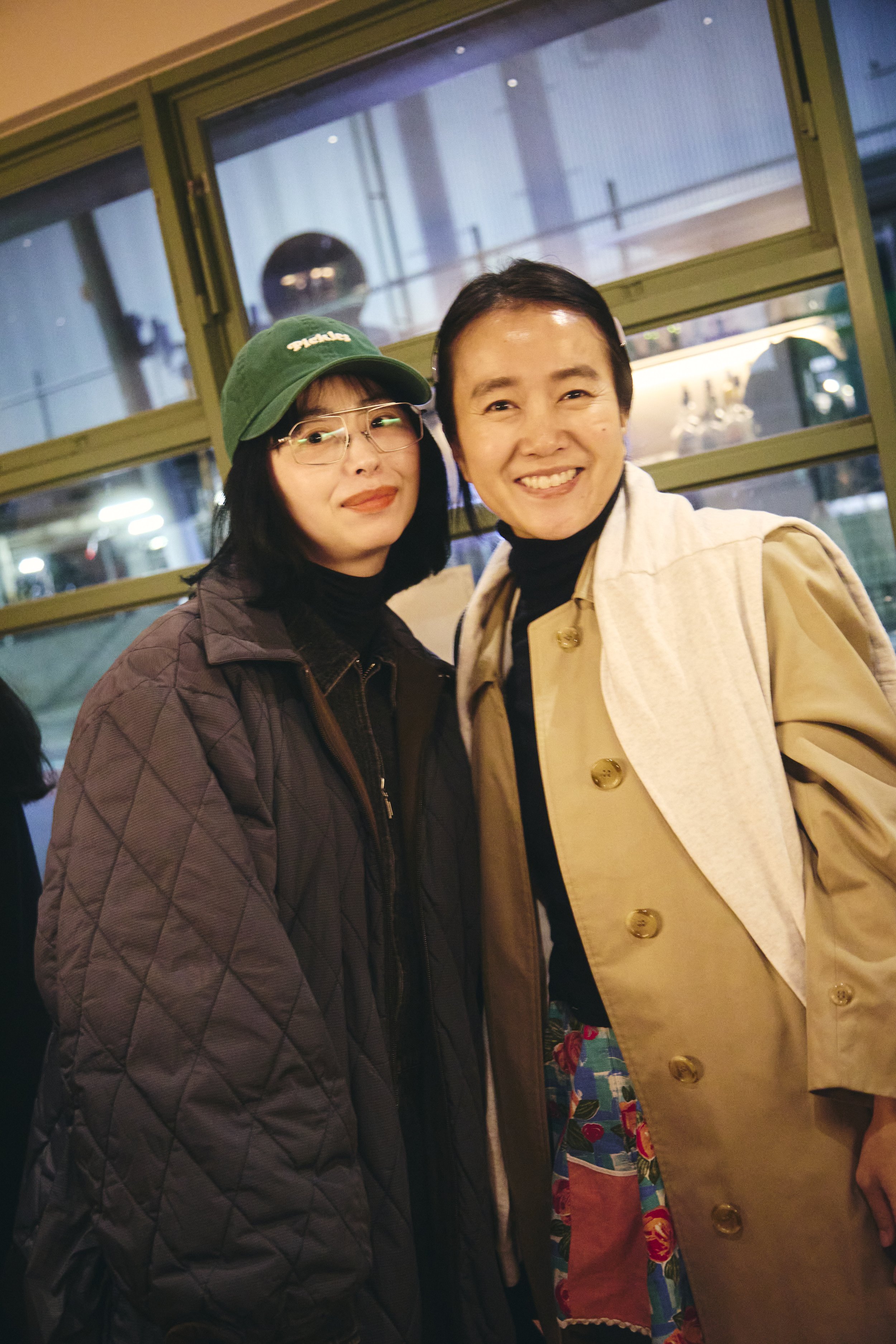 Two women standing close together indoors, smiling at the camera. The woman on the left has shoulder-length dark hair, wearing glasses, a green cap, and a dark quilted jacket. The woman on the right has short dark hair, wearing a beige coat over a bl