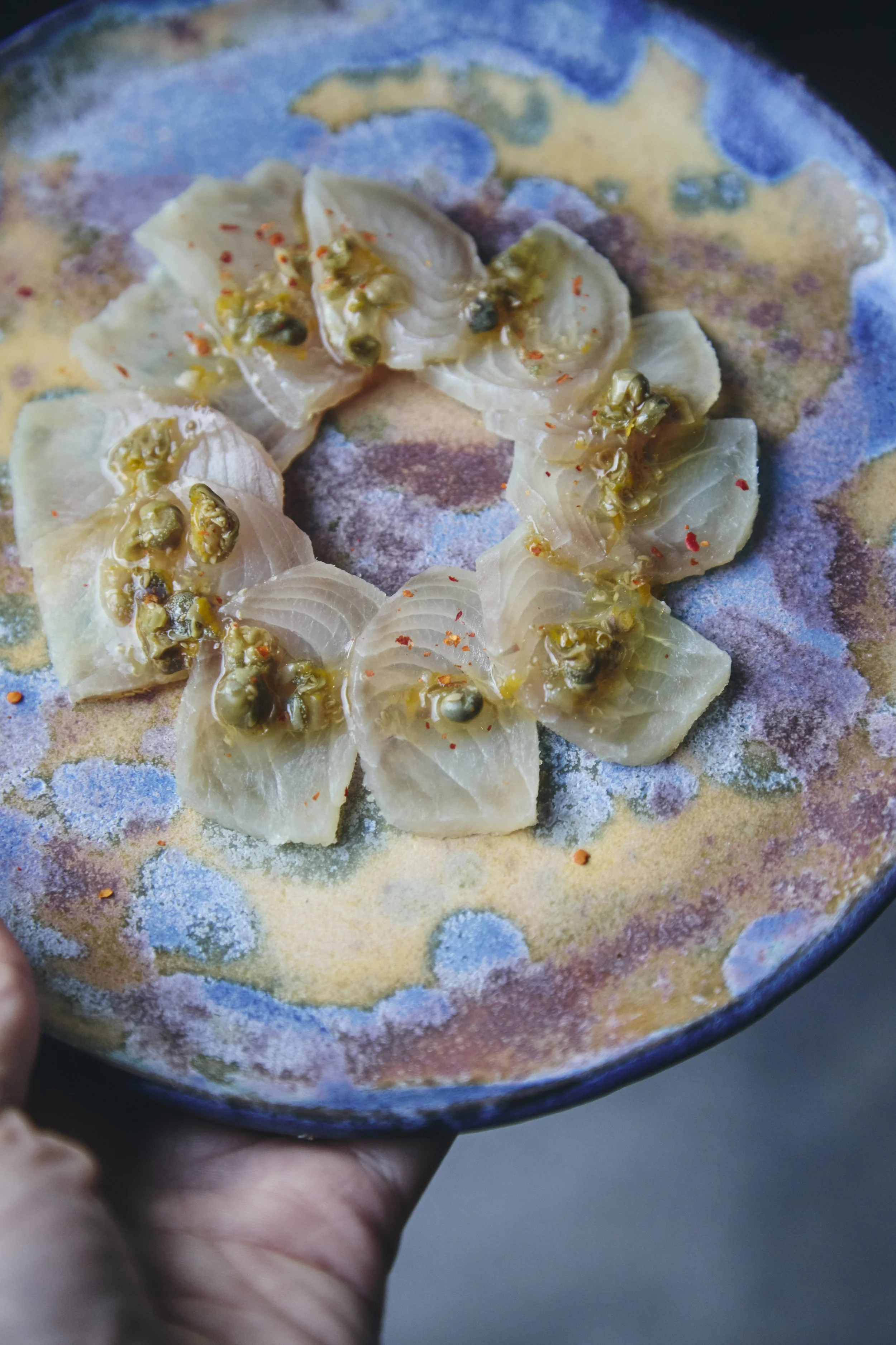 A hand holding a small decorative ceramic plate with a colorful, speckled pattern. On the plate, there are raw scallops topped with green capers and red pepper flakes.