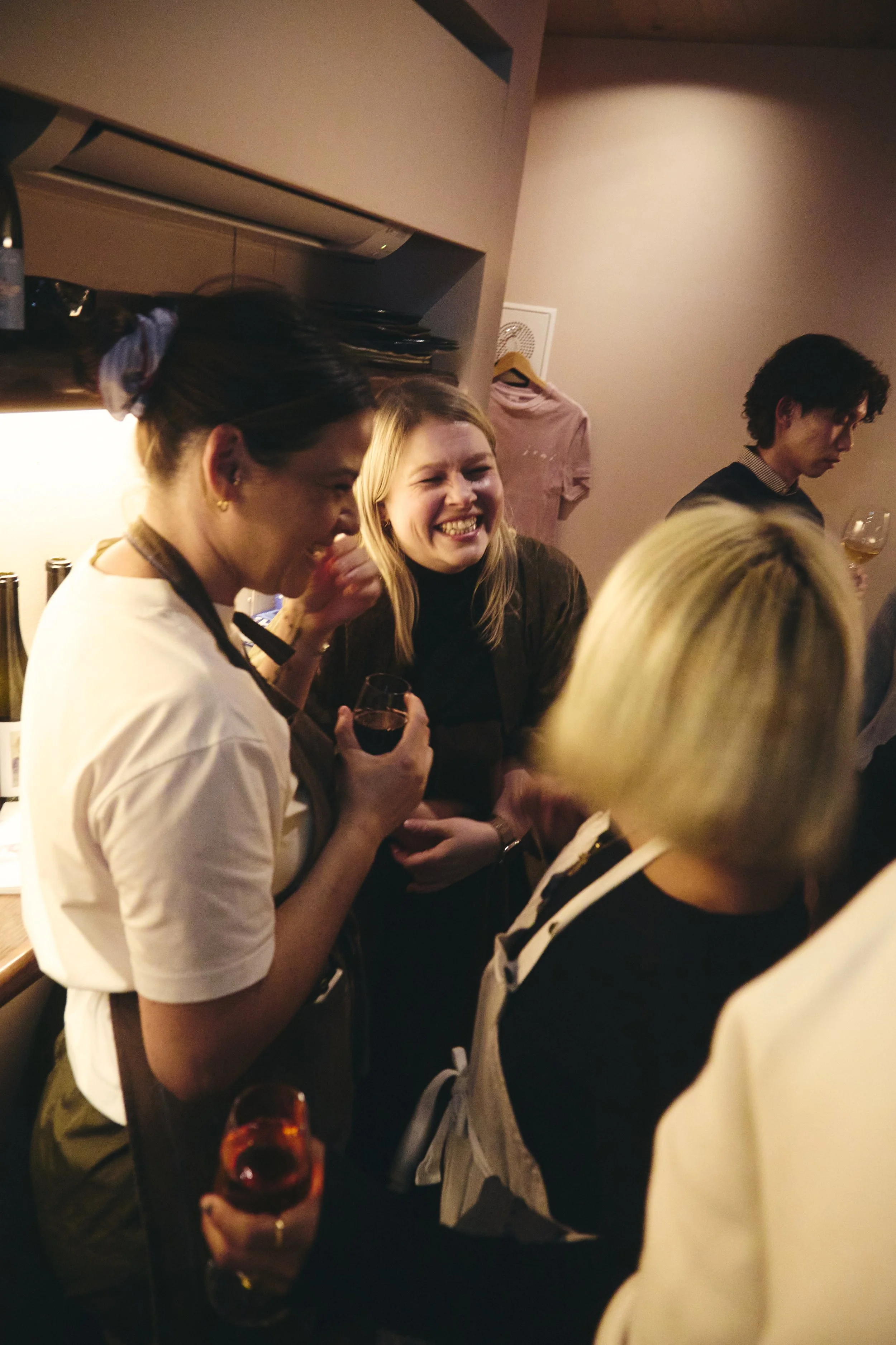 A group of people socializing indoors, with one woman laughing and holding a glass of red wine, and other individuals engaged in conversation. The setting appears cozy with warm lighting.
