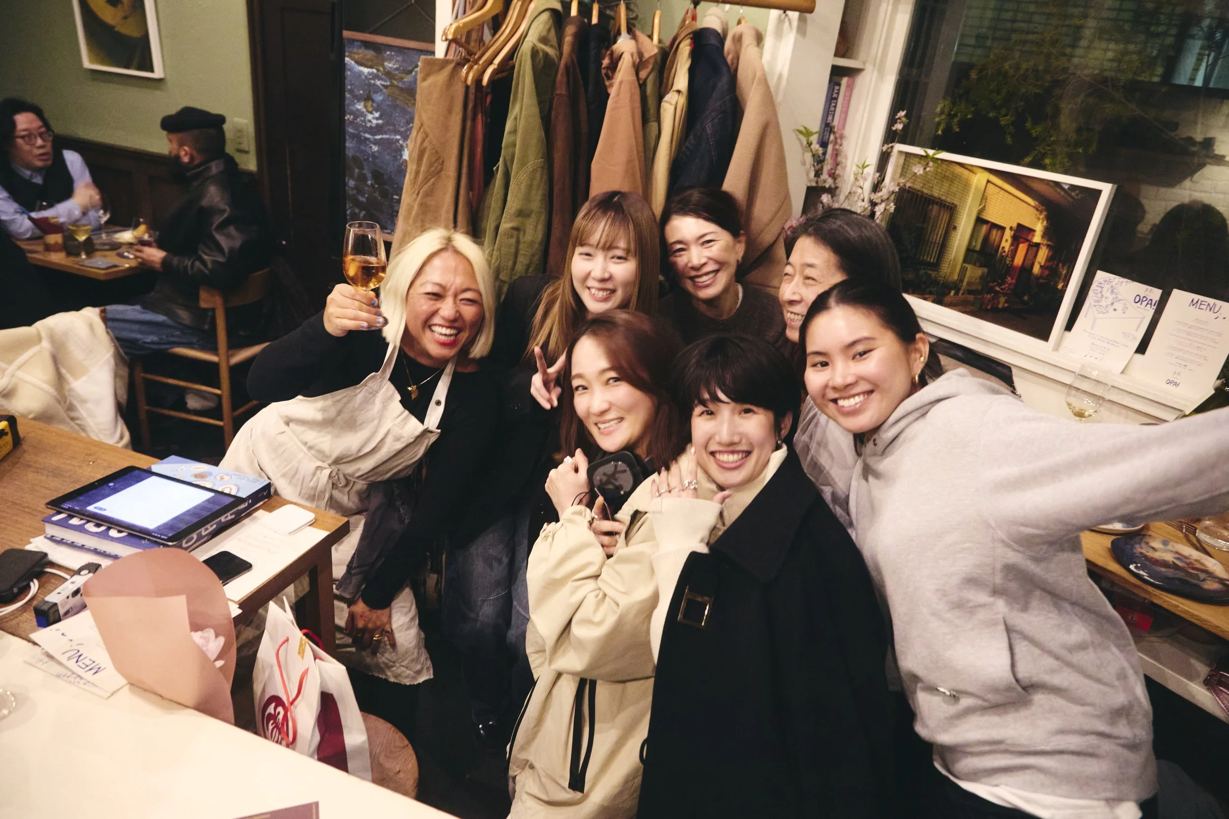 Group of six women smiling and taking a selfie at a restaurant, with jackets hanging on a rack behind them, and a table with drinks and menus in front.
