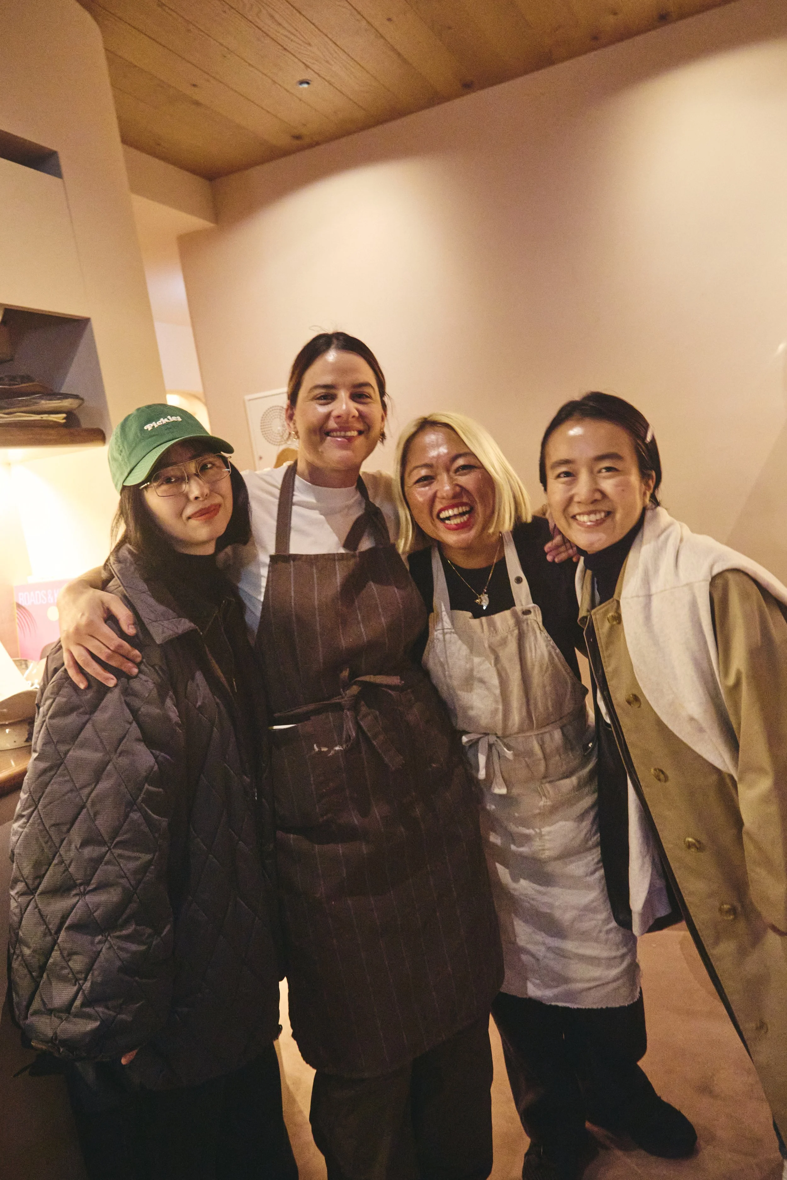 Four women standing close together and smiling in a warmly lit indoor space, with one woman wearing a green baseball cap and glasses, another in a dark apron, and others wearing aprons and casual clothing.