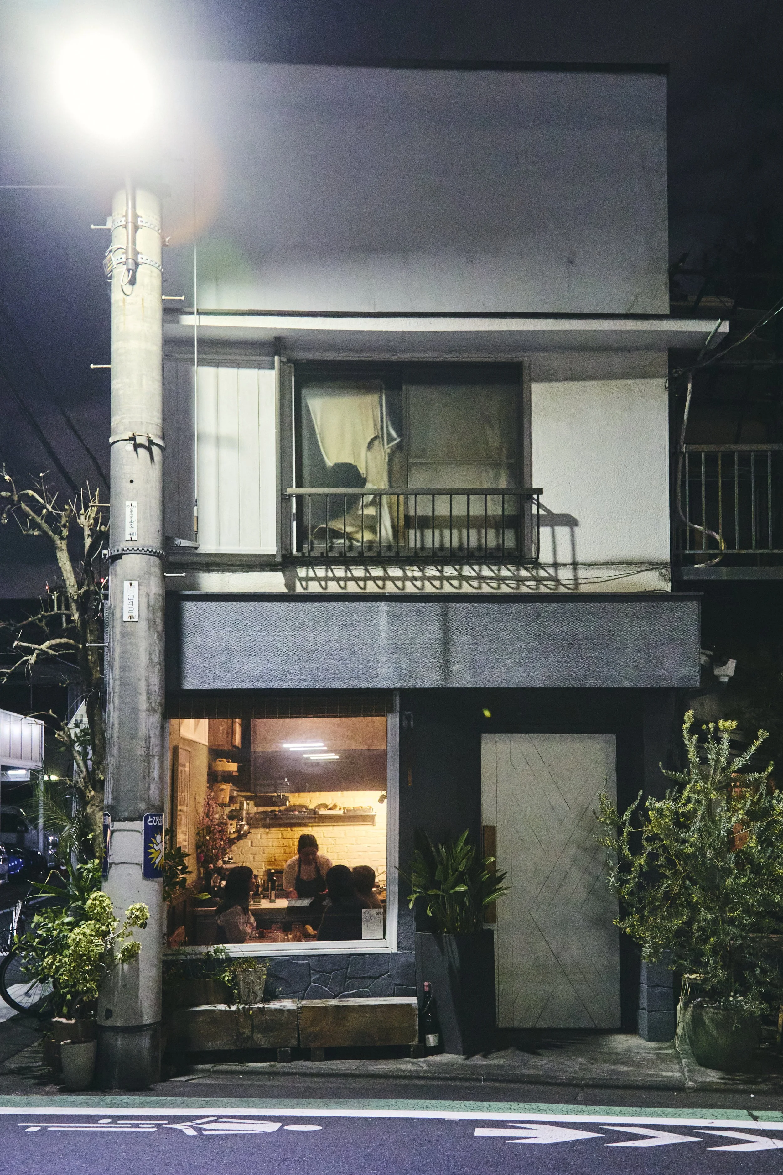 A multi-story building at night, with a restaurant on the ground floor occupied by several people dining, and a small balcony on the second floor. The building is lit with warm lighting inside, and a tall street lamp is visible at the street level. T