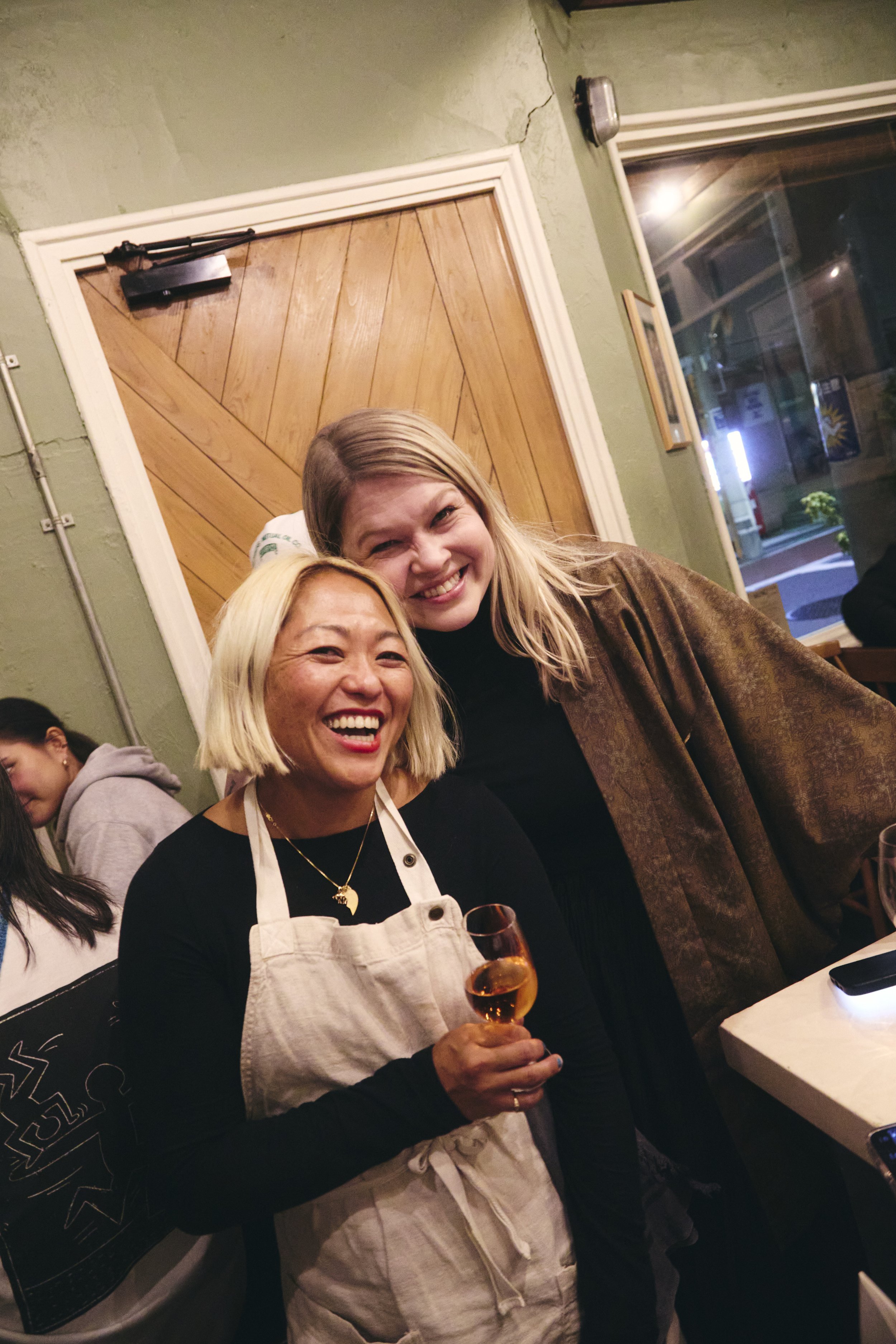 Two women smiling, one with short blonde hair holding a glass of rosé wine, the other with long blonde hair in a brown coat, inside a restaurant at night.