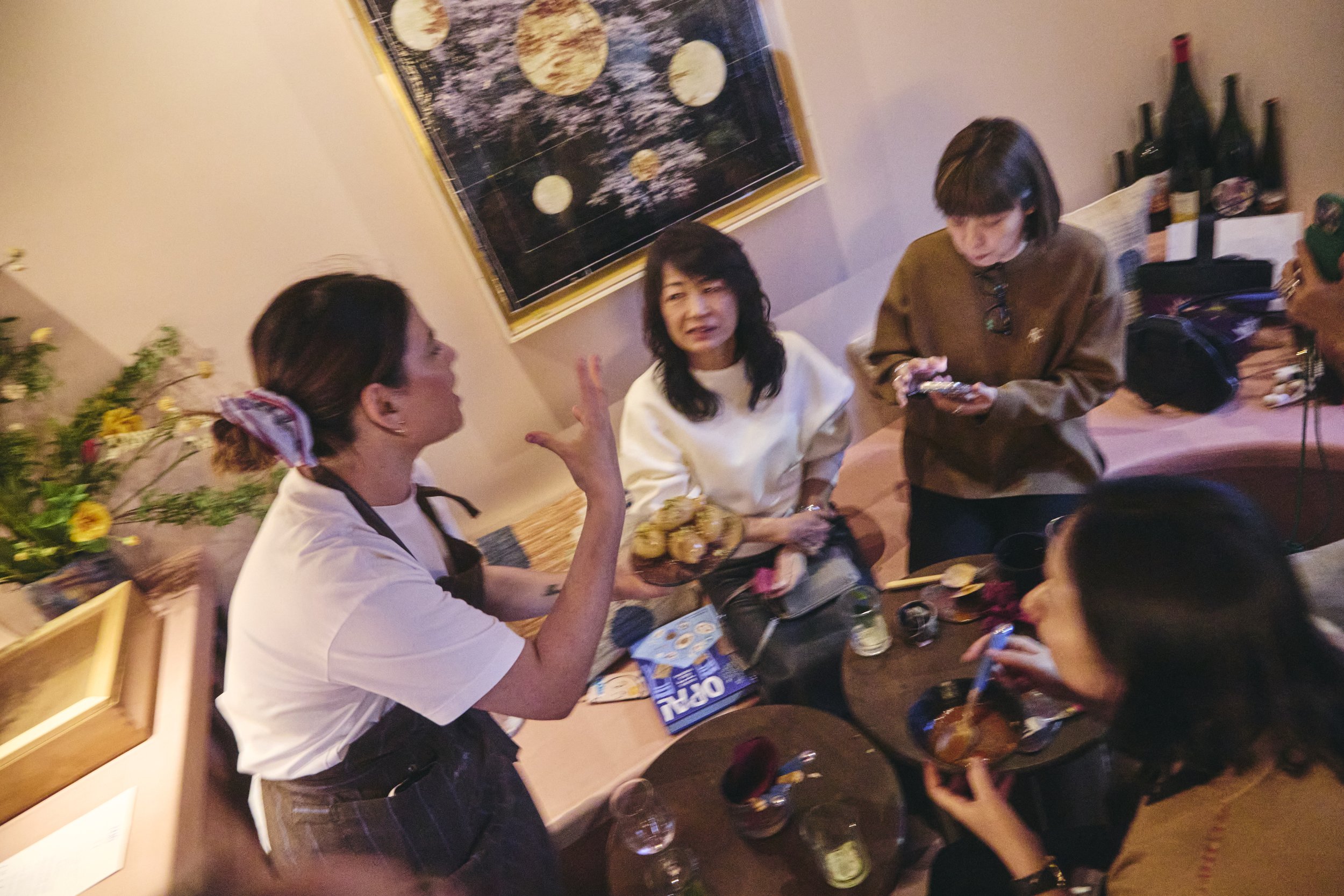 Group of people talking at a party or gathering with bottles of wine on the table and a colorful artwork on the wall behind them.