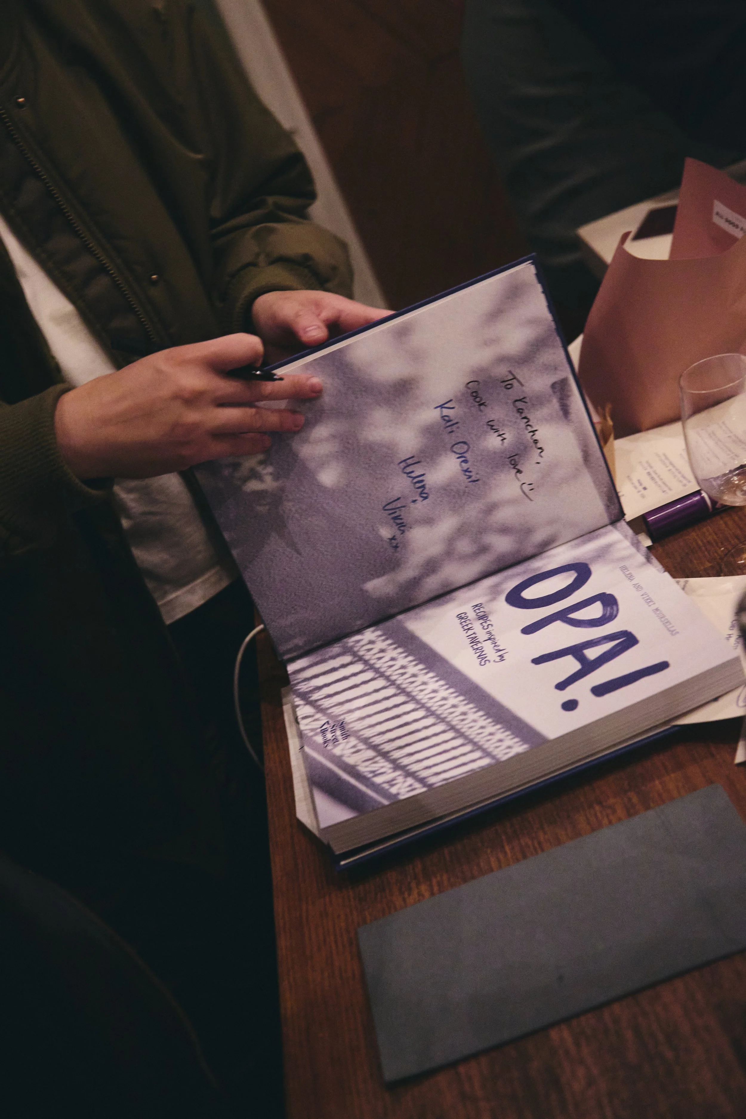 Close-up of a person holding an open menu at a restaurant table, with the menu showing the words "To Vegetarians, Cook with Love" and handwritten notes, surrounded by a wine glass and paper materials.