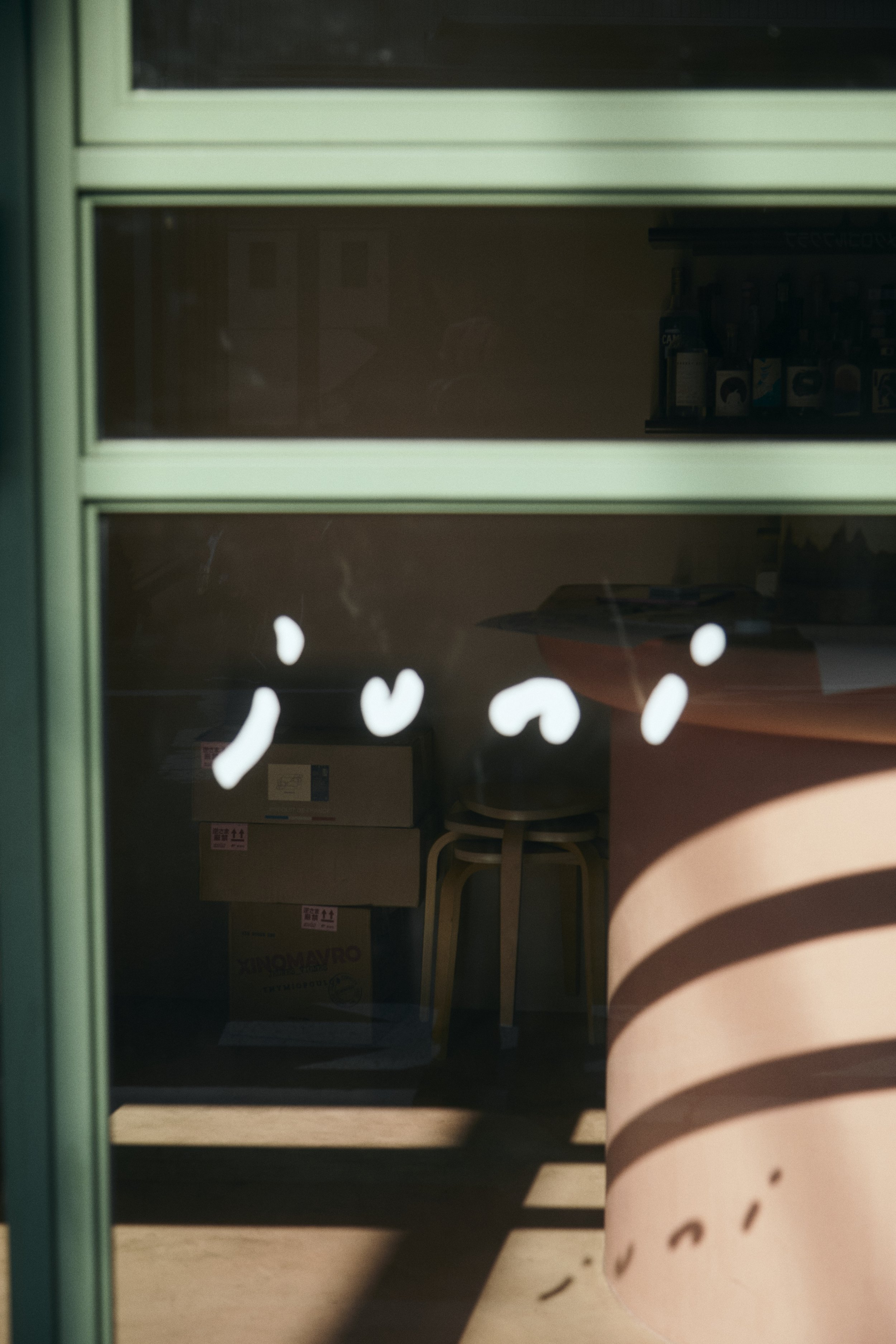 View through a glass window showing cardboard boxes stacked inside a room and part of a wooden table with a chair. Reflection of a person's hand and face on the glass.