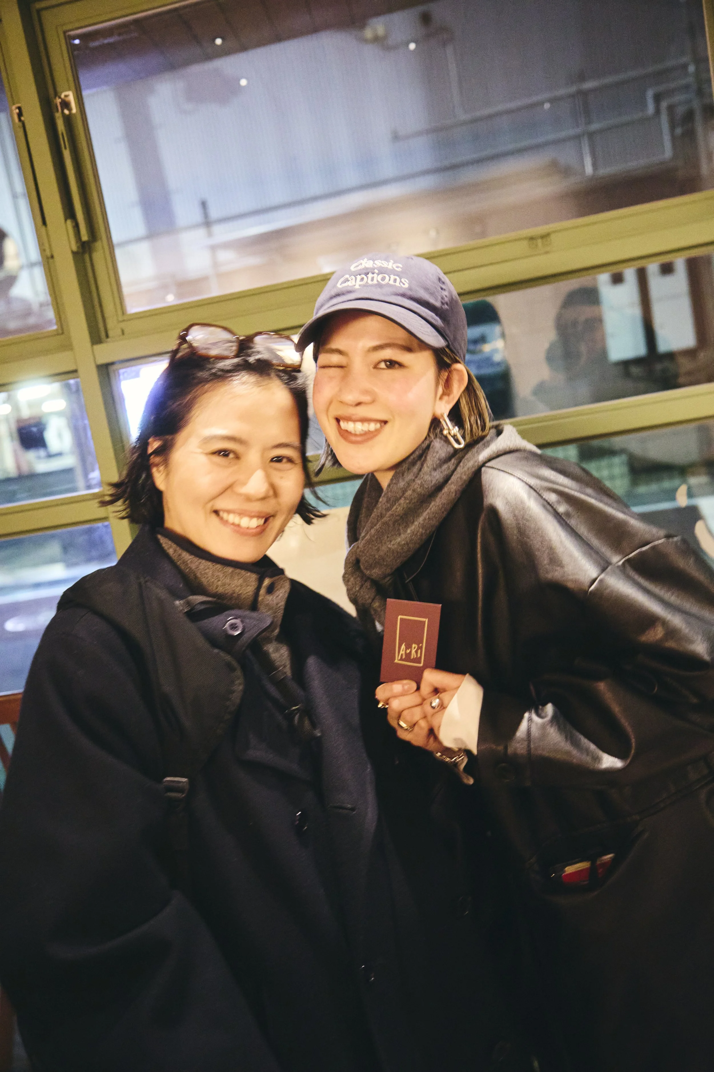 Two women smiling at the camera, one holding a red card with the letters 'A-R-I', standing in front of a window.