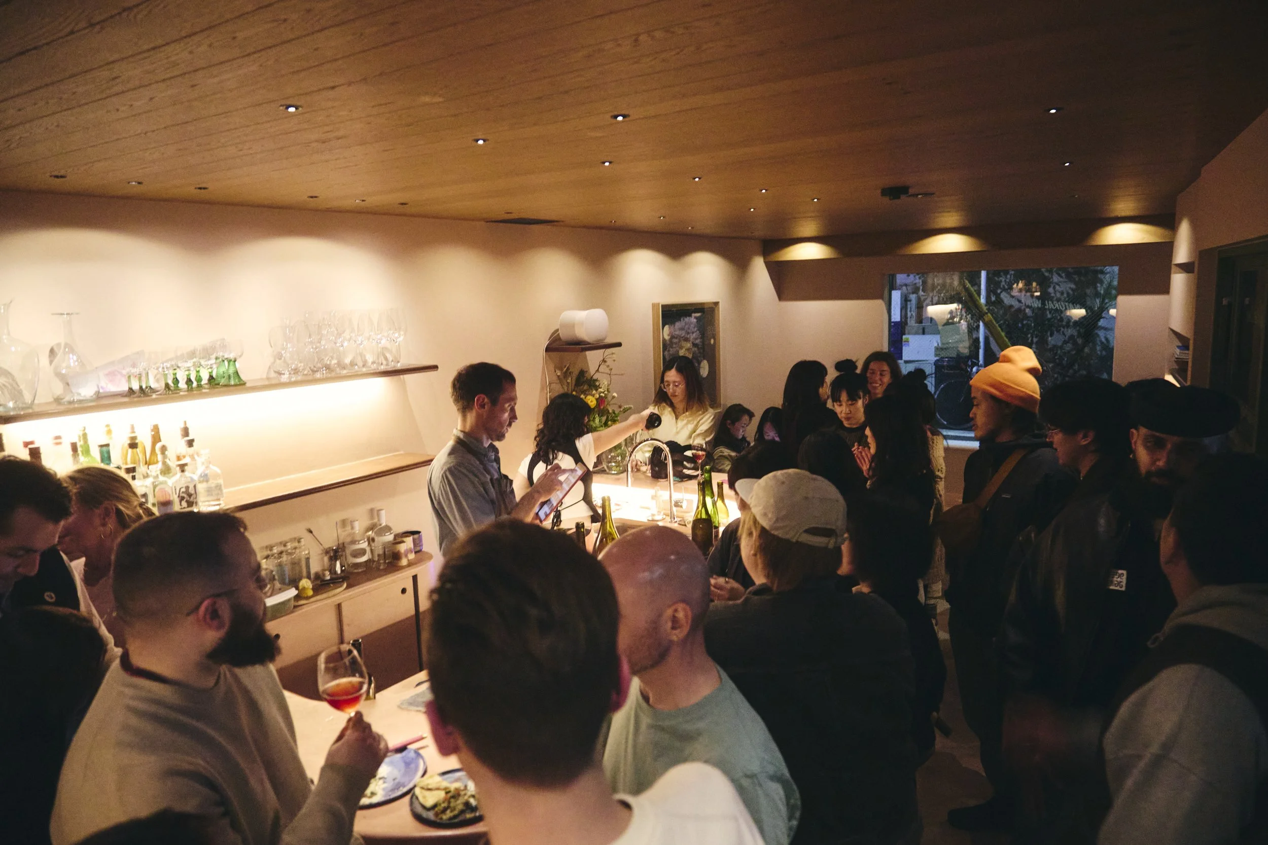 A crowded indoor bar or restaurant with people socializing, some holding drinks. There are shelves with glassware and bottles behind the bar, and a window showing plants outside. The setting has warm lighting and a wooden ceiling.