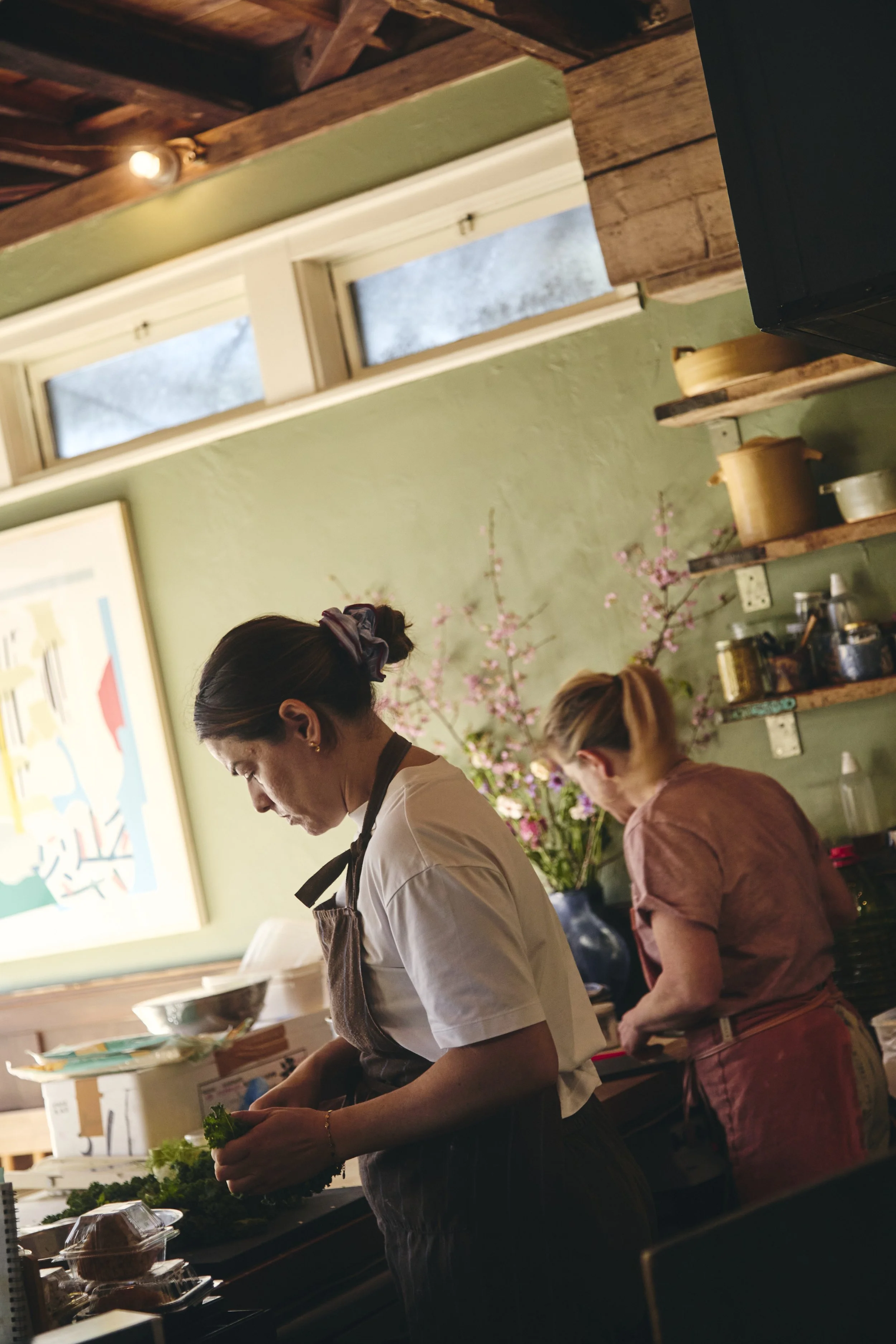 Two women preparing food in a cozy kitchen with green walls, wooden shelves, and pink flowers in the background.