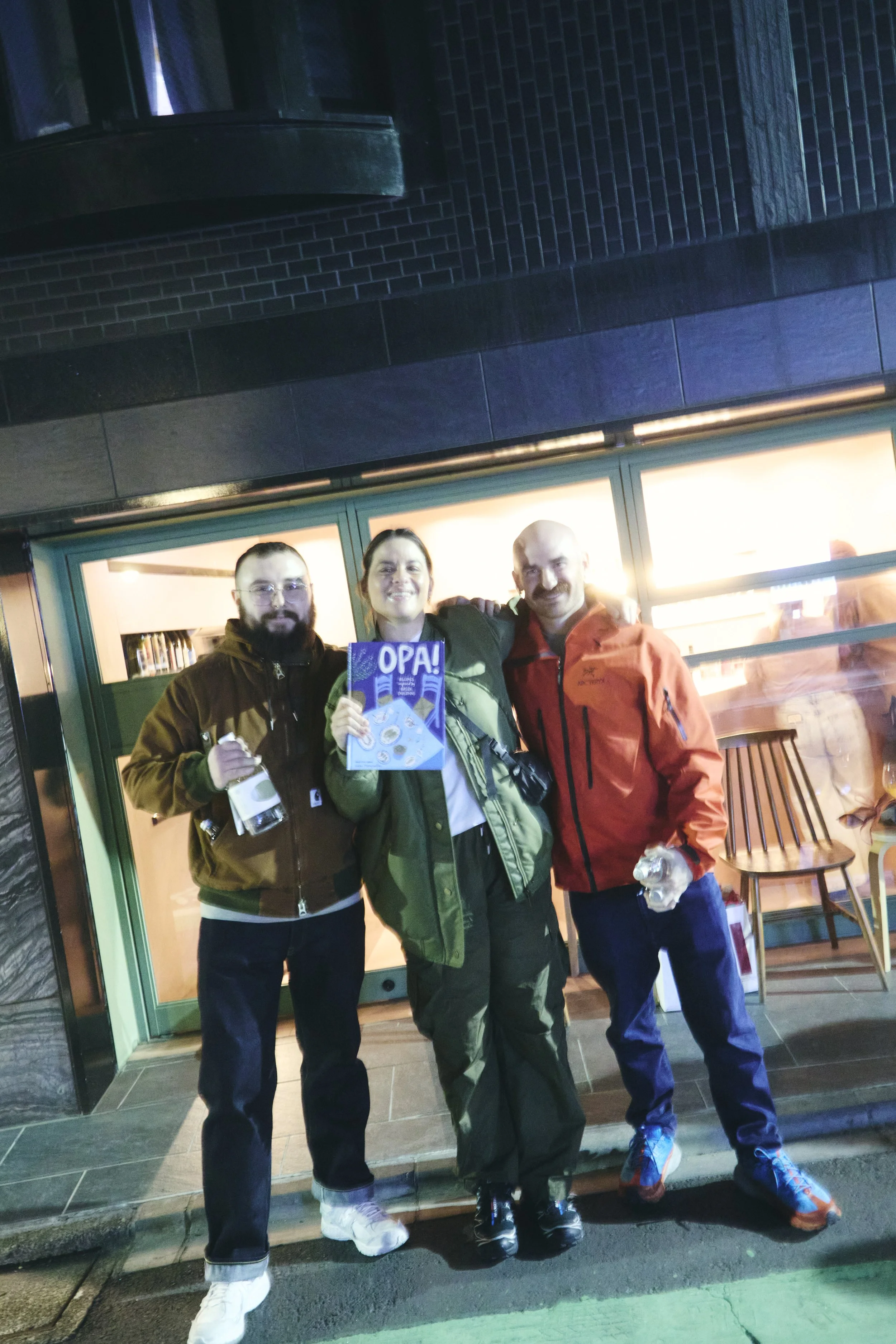 Three people are standing together outside a building at night. The person in the middle is holding a colorful booklet that says "OPA!" and smiling. The person on the left is holding a water bottle, and the person on the right is holding a plastic bo