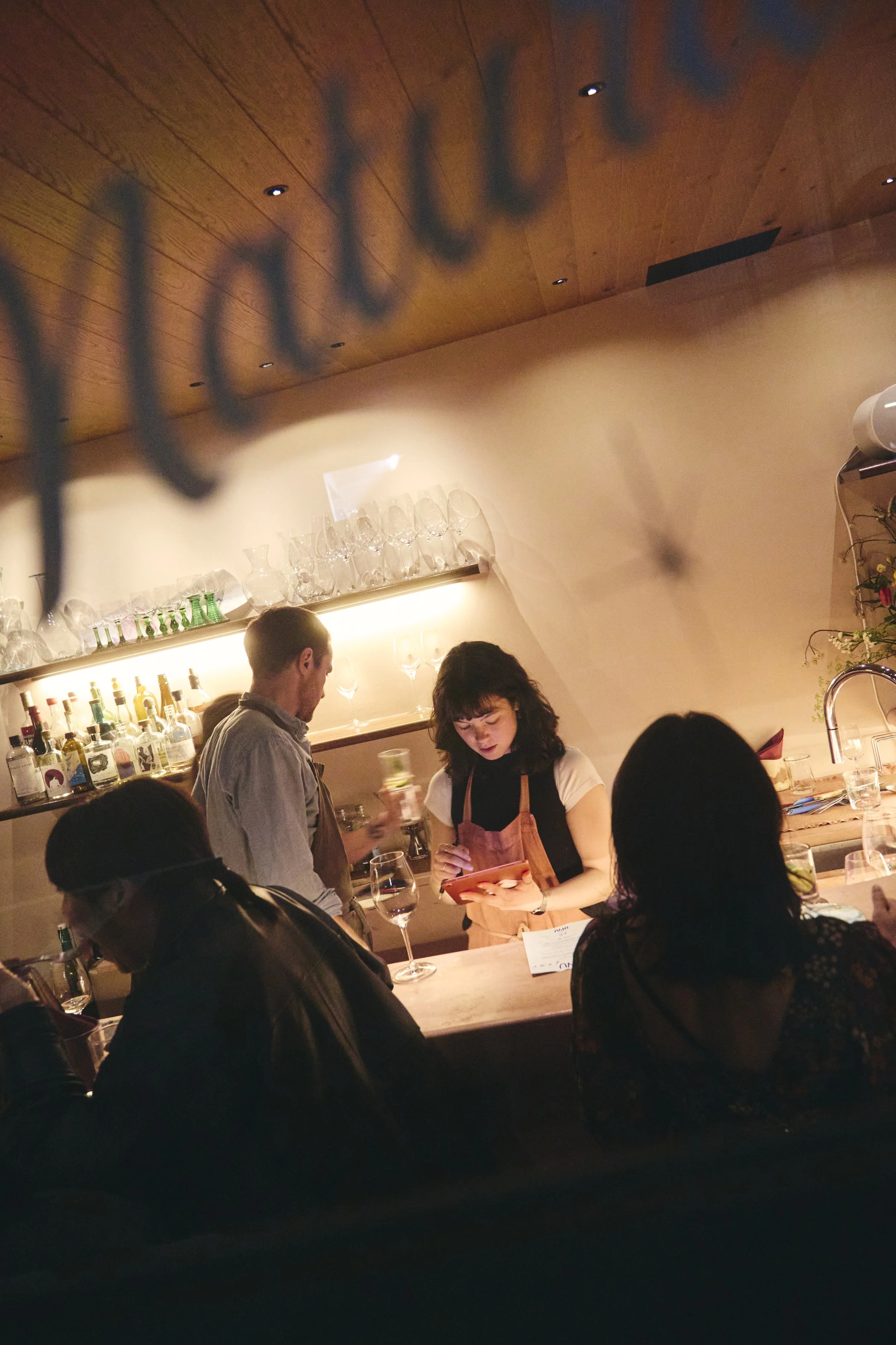 A woman in an apron holding a tablet at a bar with several people around, bar shelves with bottles and glasses on the wall behind her.