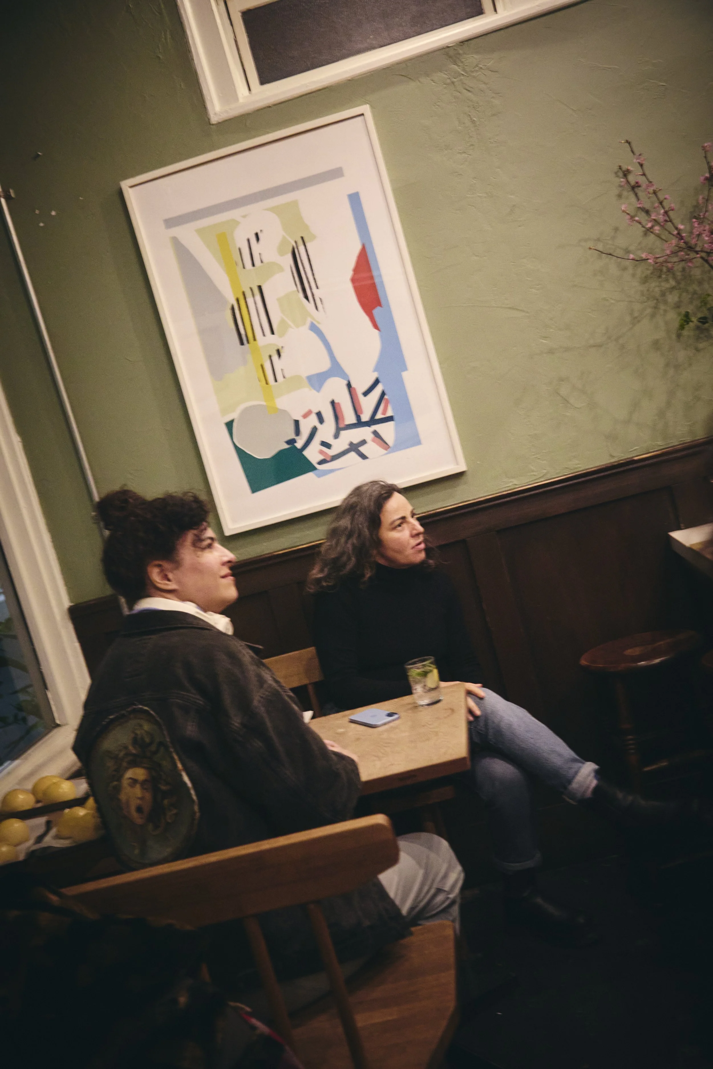 People sitting at tables in a restaurant, with a large abstract art piece on the green wall behind them and a window above, a glass of water and a smartphone are on the table.