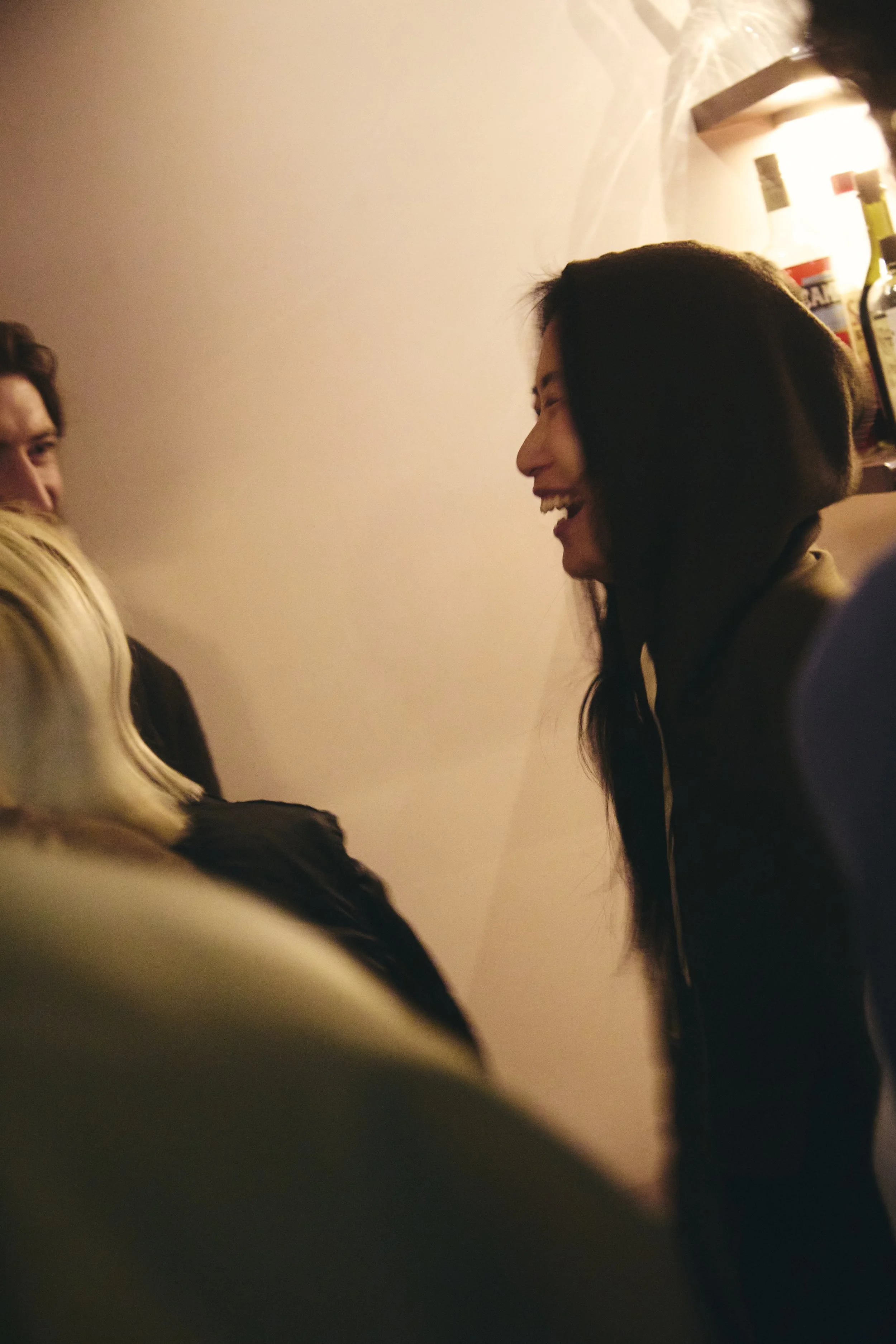 People are socializing in a room with a plain wall, and a woman with long dark hair is laughing, engaging with others.