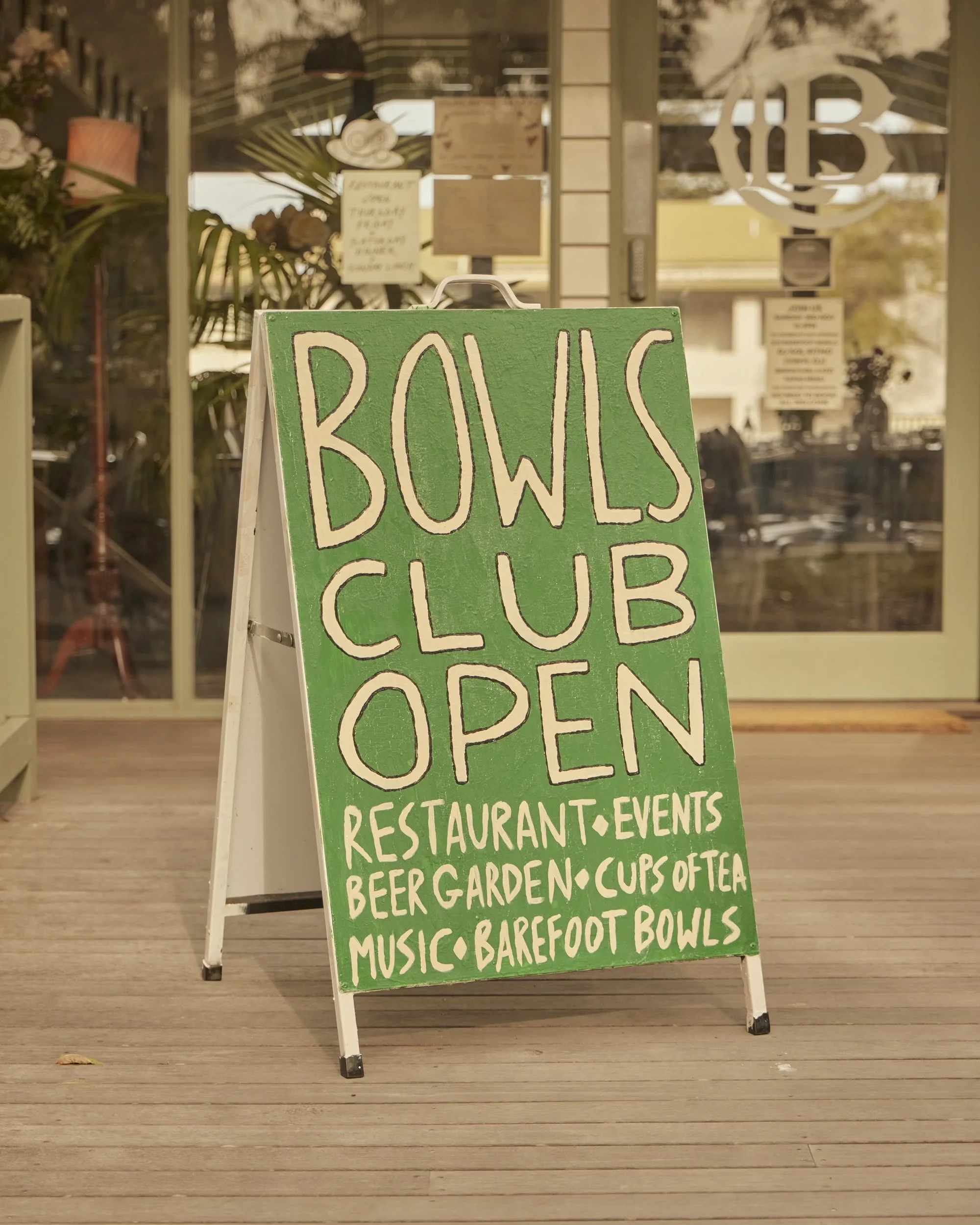A green and white sidewalk sign announcing that Bowls Club is open, with a list of offerings including restaurant, events, beer garden, cups of tea, and barefoot bowls. The background shows a glass door and windows with blurred indoor details and ref