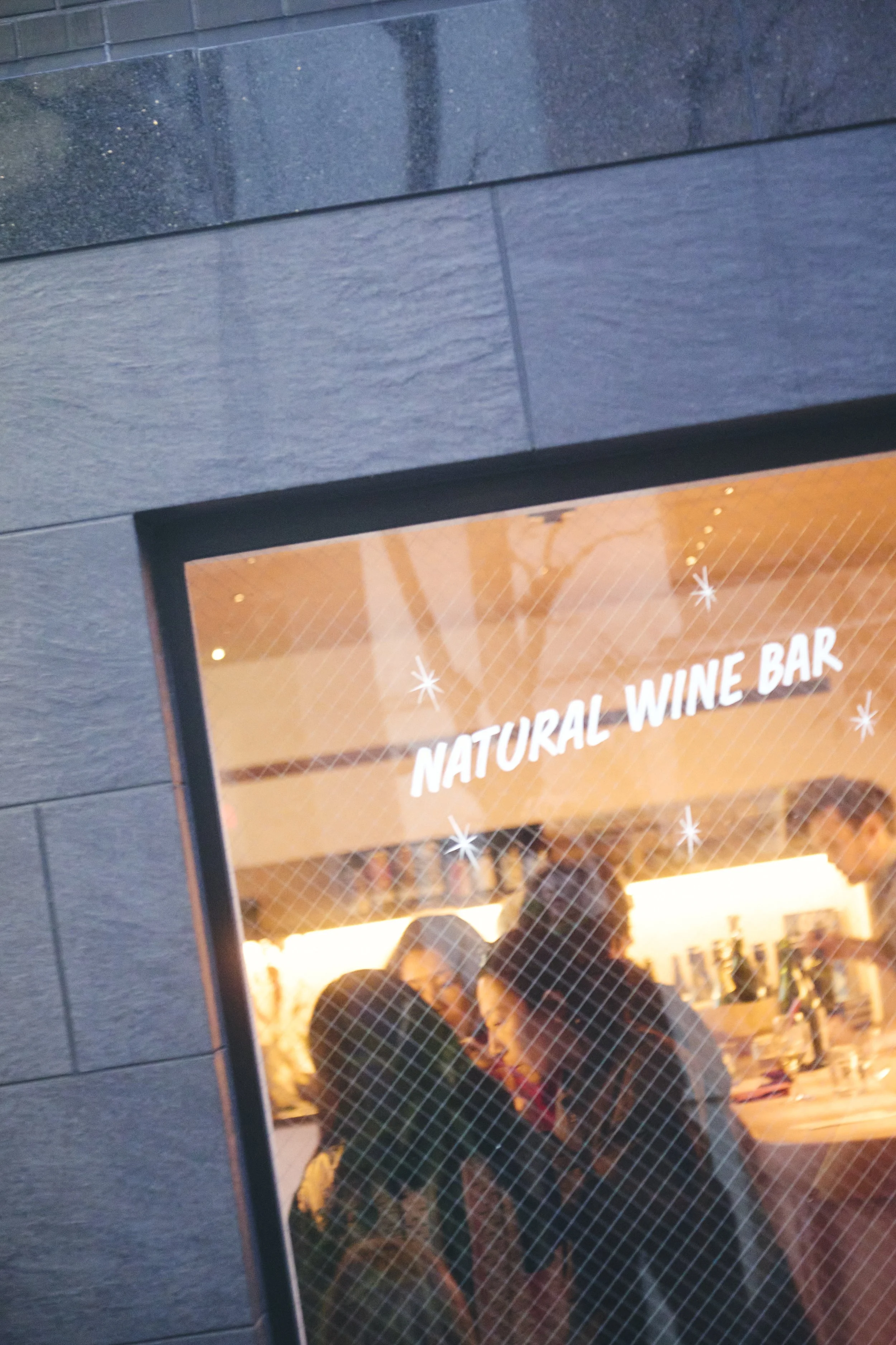 People inside a restaurant or bar, viewed through a glass window with a wire mesh pattern, with a sign above that reads 'NATURAL WINE BAR'.